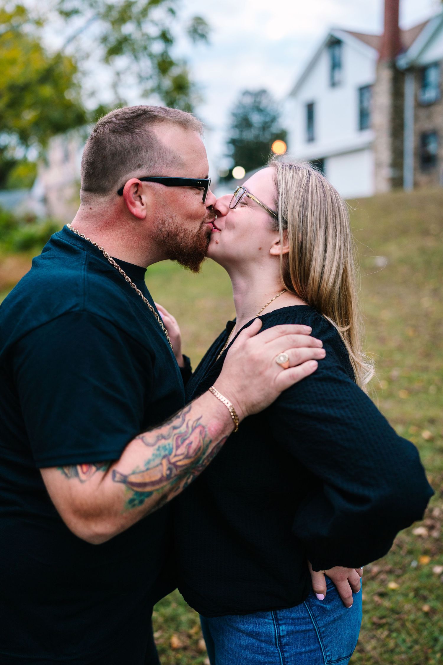 Philly wedding photographer tattooed man and blonde woman sharing a romantic kiss outdoors, both wearing black tops in a suburban setting.