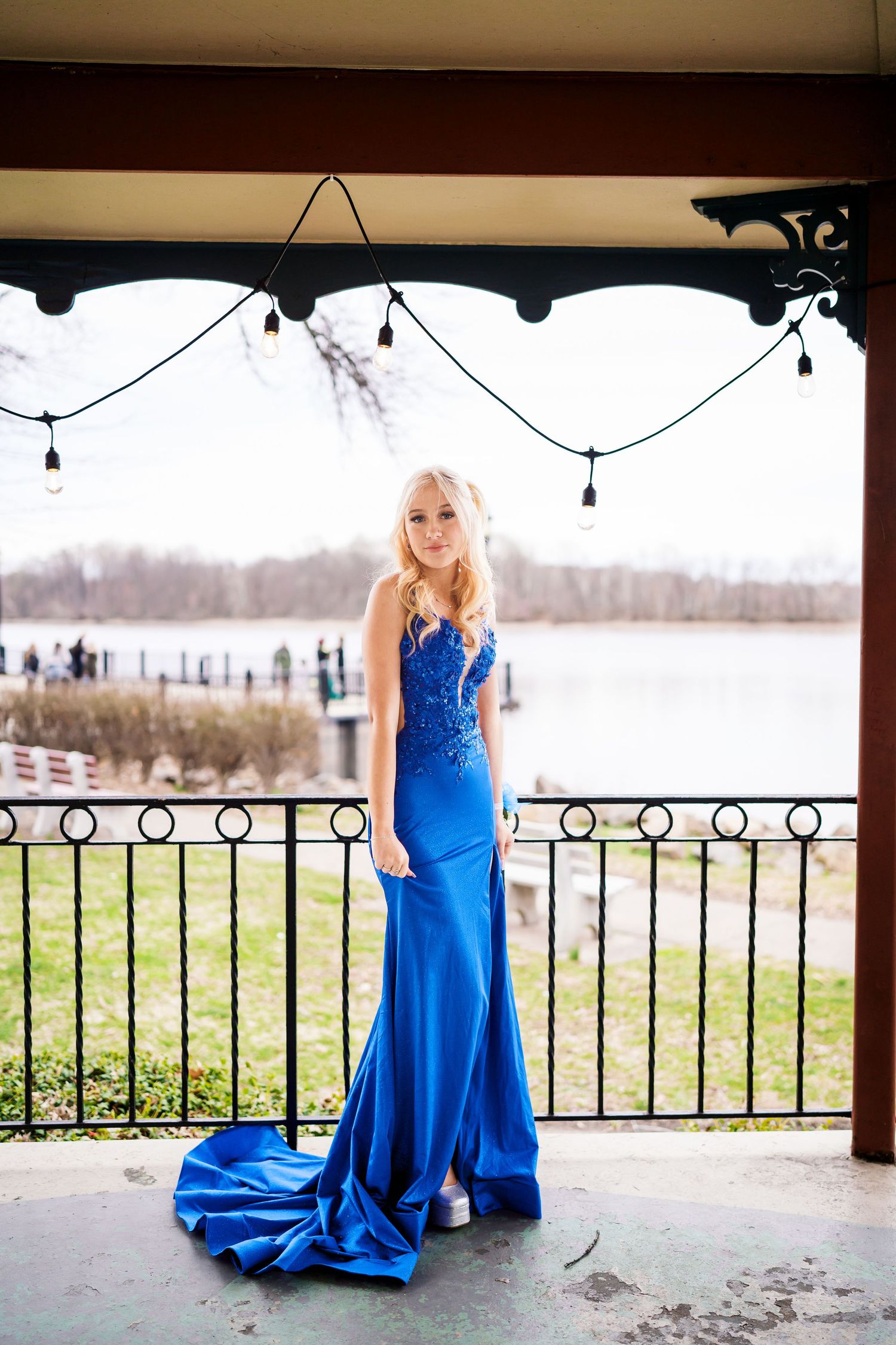 Young woman in elegant blue satin mermaid gown with train poses on outdoor balcony overlooking a lake.