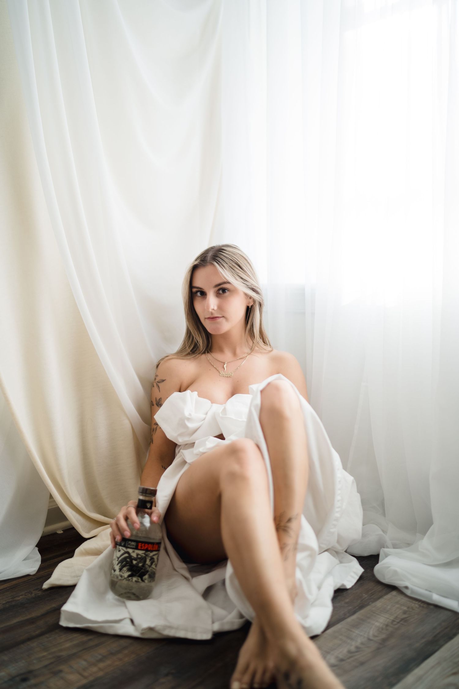 Young blonde woman in white fabric sitting on floor holding a bottle near sheer curtains in soft natural light.
