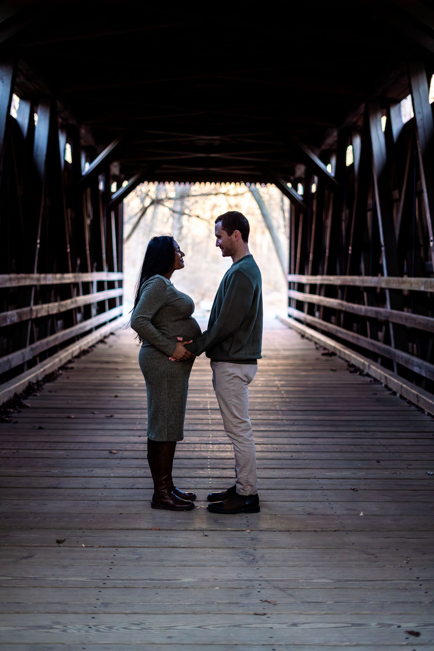 Couple standing face-to-face on a wooden covered bridge, woman cradling pregnant belly in a maternity photo.