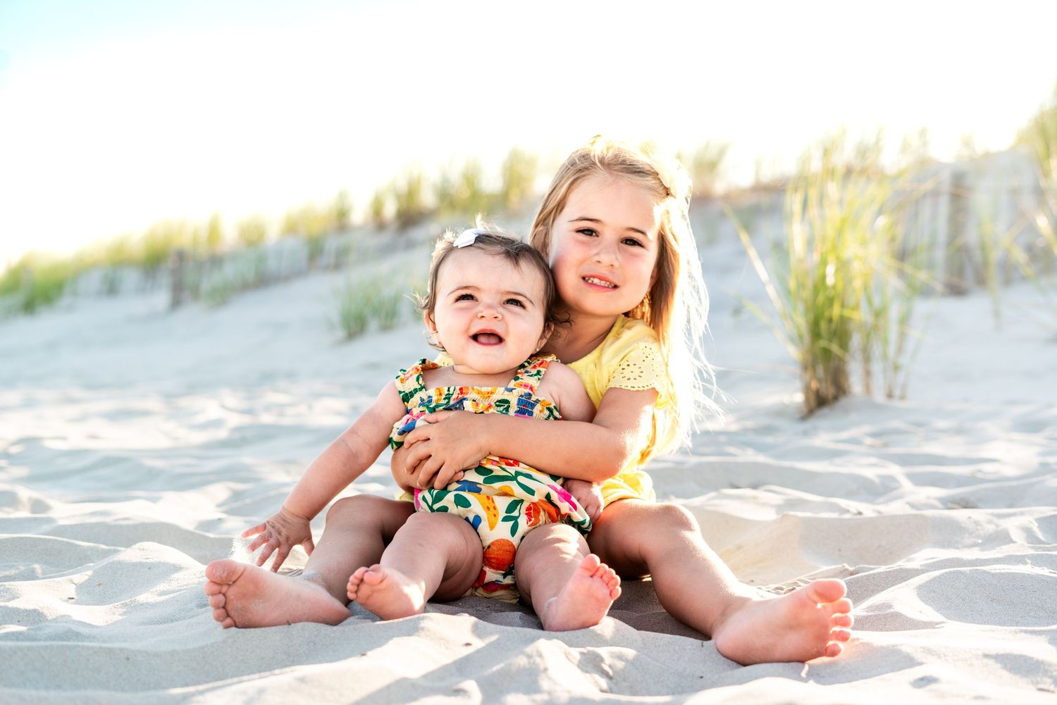Two happy sisters sitting on sandy beach, older girl hugging baby in colorful swimsuit, sunny dunes background.