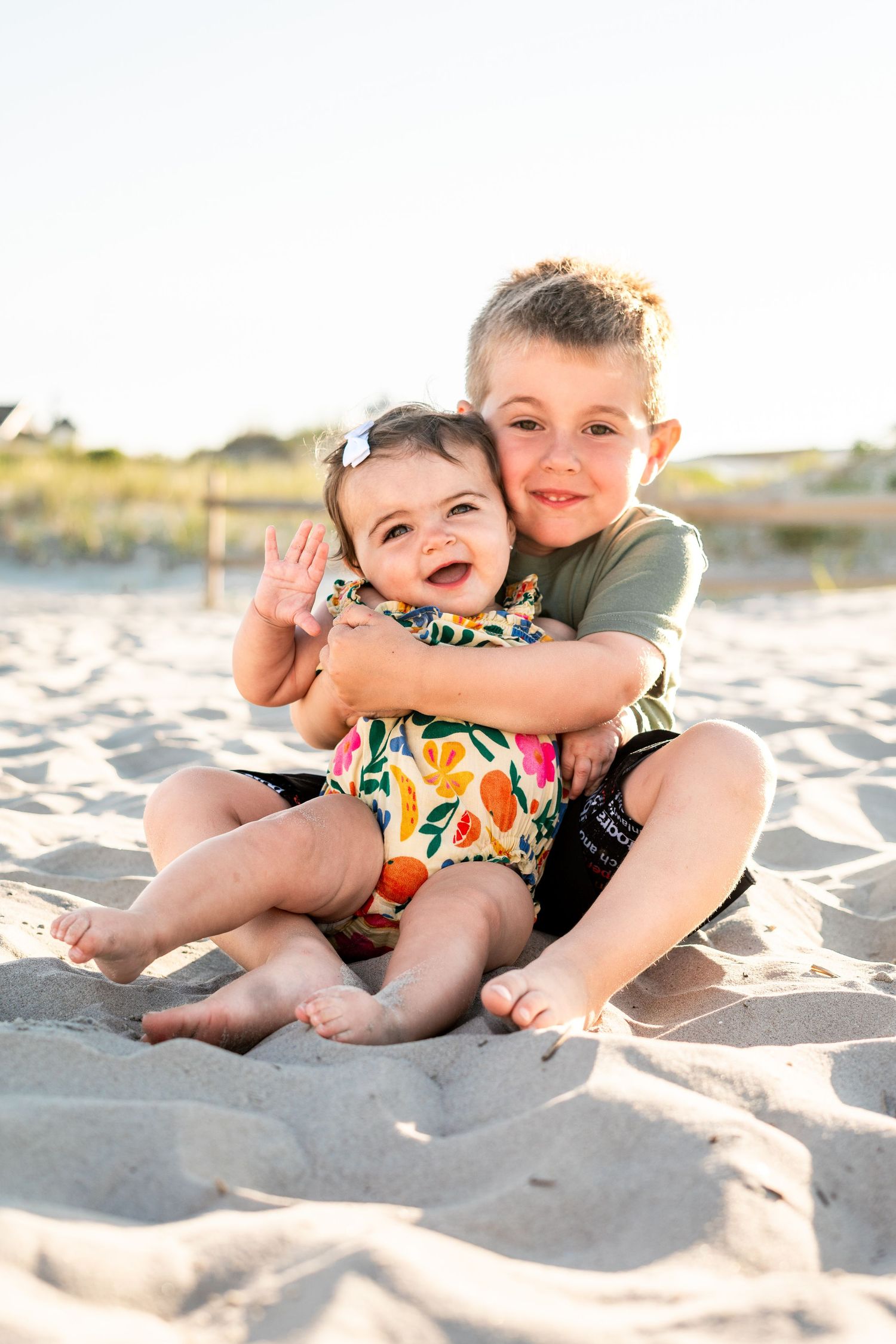 Two young siblings hugging and smiling on a sandy beach, baby in floral outfit waving, older boy in green shirt.