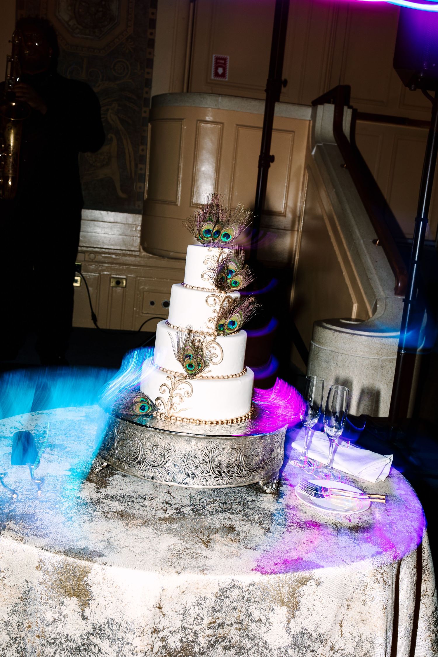 Elegant four-tier white wedding cake adorned with peacock feathers on a silver stand, illuminated by colorful blue and pink lights.