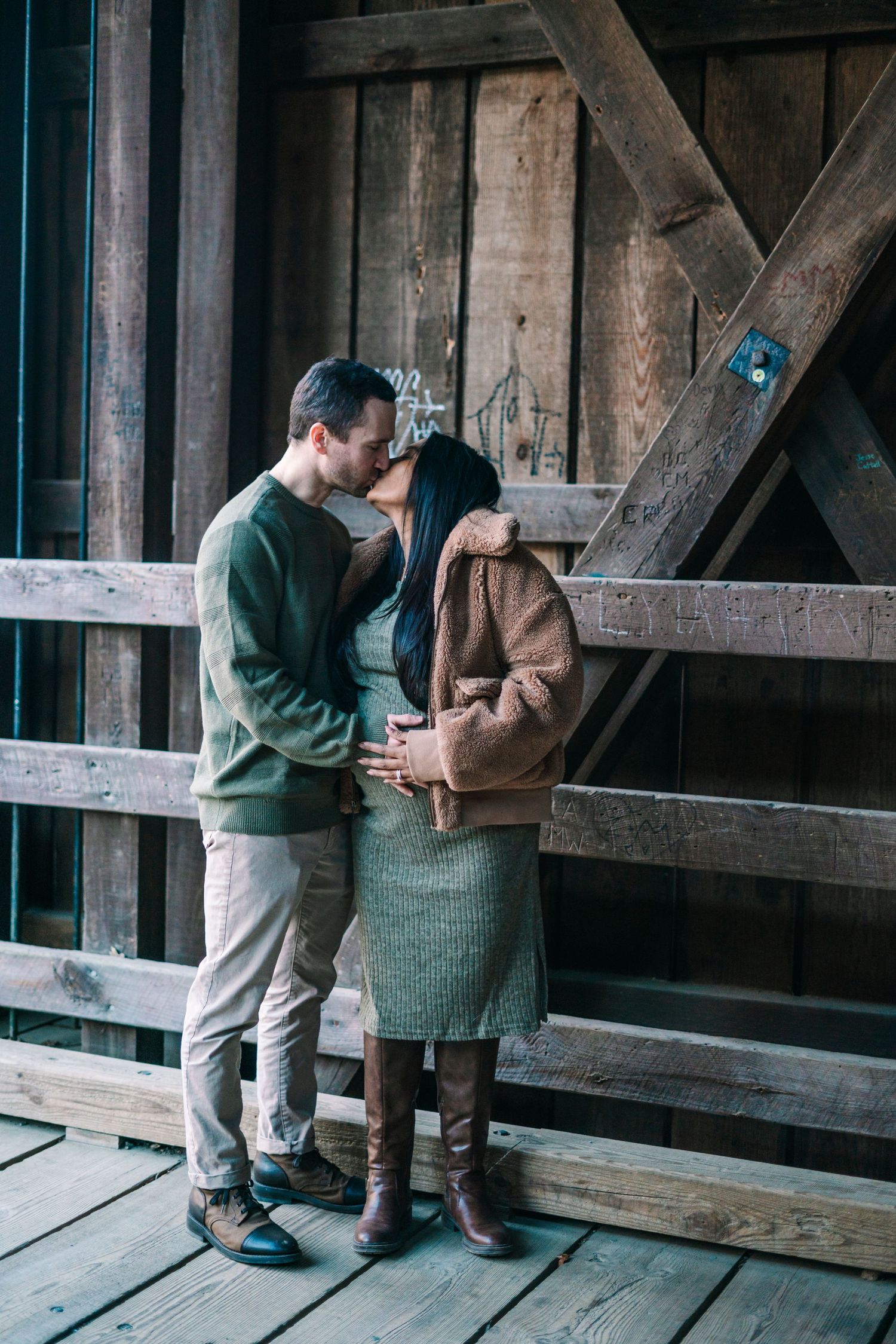 Couple sharing a romantic kiss on a rustic wooden bridge, dressed in cozy autumn outfits of green and brown tones.