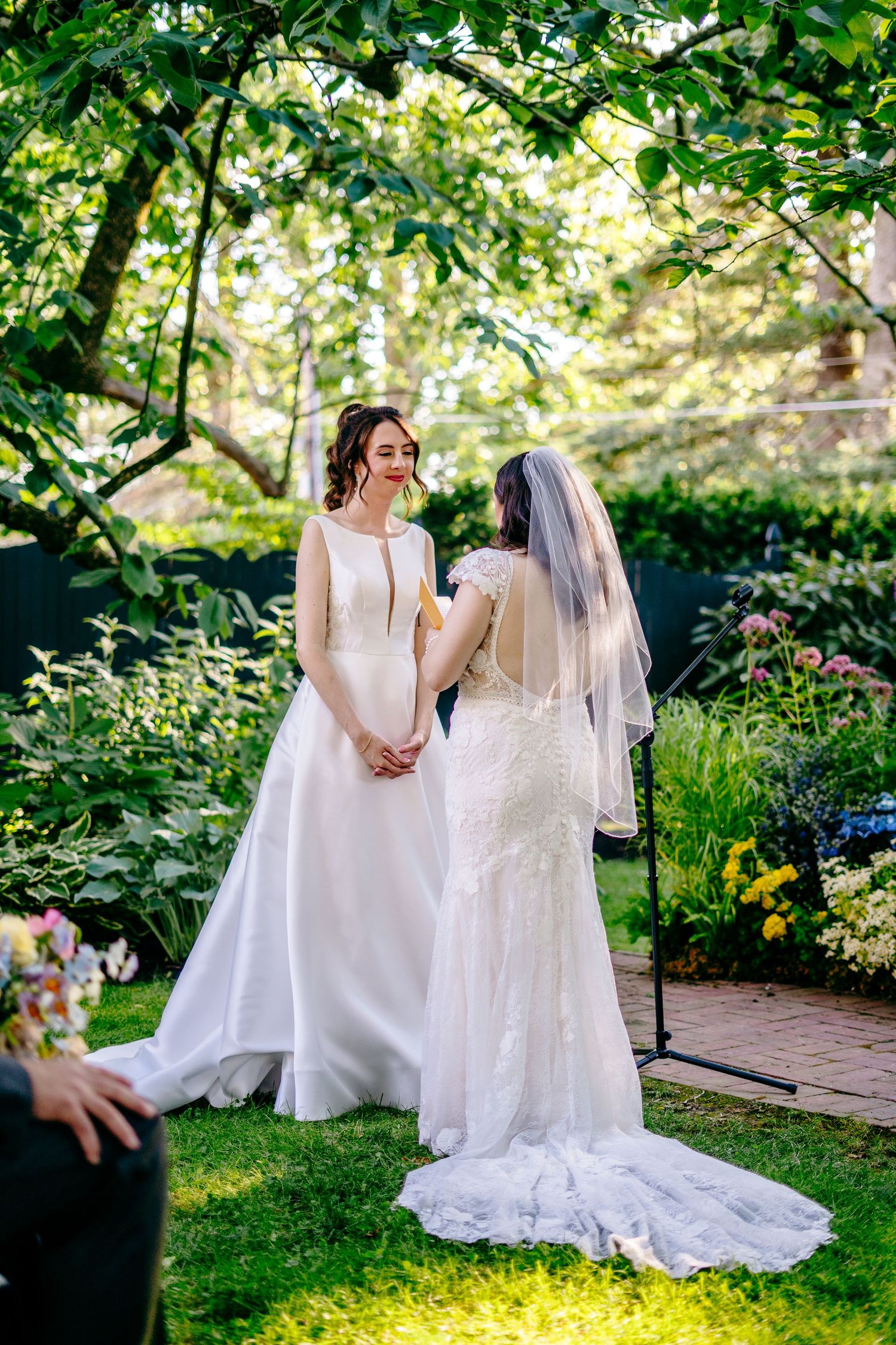 Fernbrook farms Philly wedding pTwo brides in white wedding gowns hold hands during an outdoor garden ceremony surrounded by lush greenery.