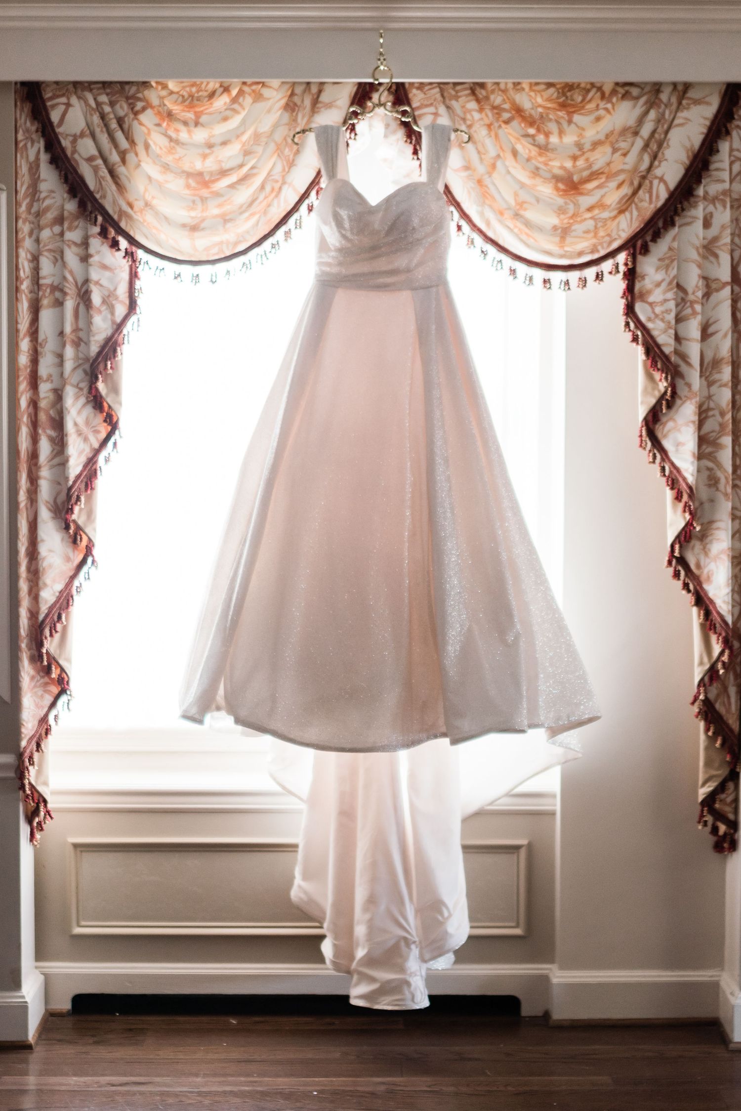 Elegant white wedding dress hanging in front of a window with ornate floral curtains in a bright room Hotel DuPont wedding Philadelphia phot