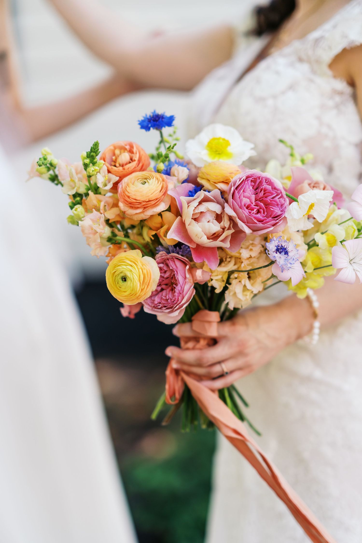 Fernbrook farms Bride holding a vibrant wedding bouquet with pink, orange, and yellow ranunculus, blue cornflowers, and peach ribbons.