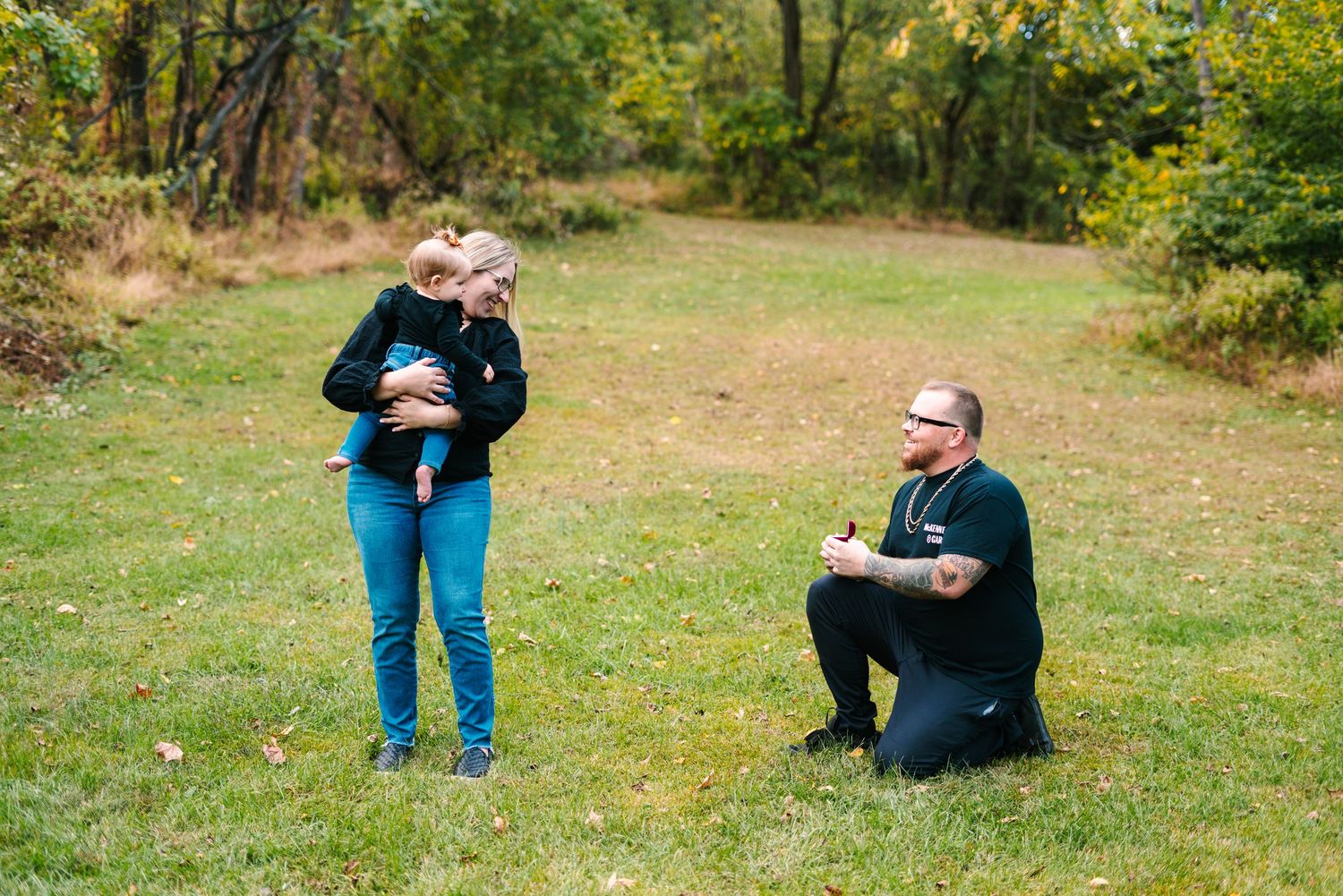 philadelphia wedding photography  A woman holding a baby talks with a tattooed man sitting on grass in an autumn park setting.