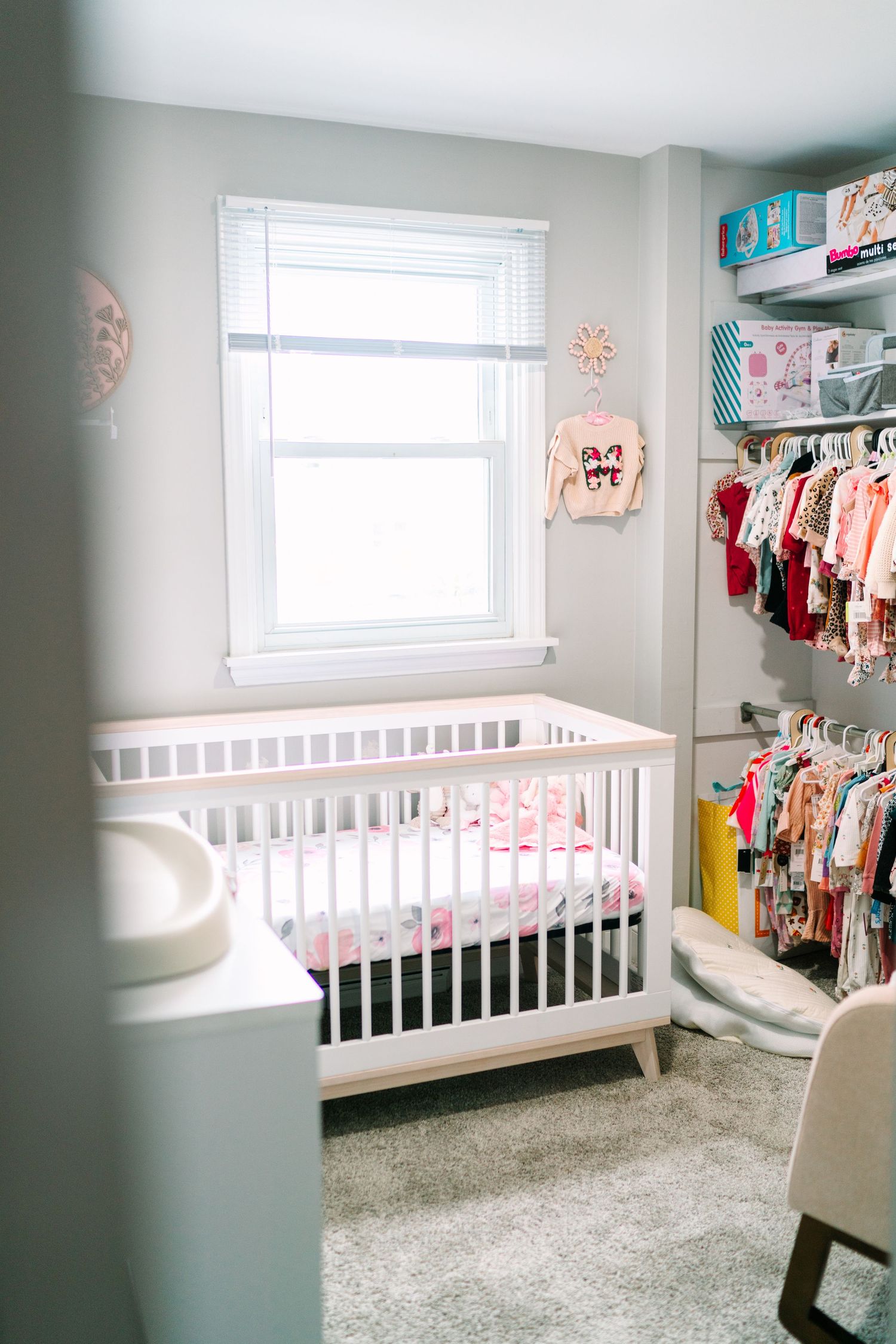 White baby crib in a cozy nursery with gray walls, carpet flooring, and colorful baby clothes on a rack.