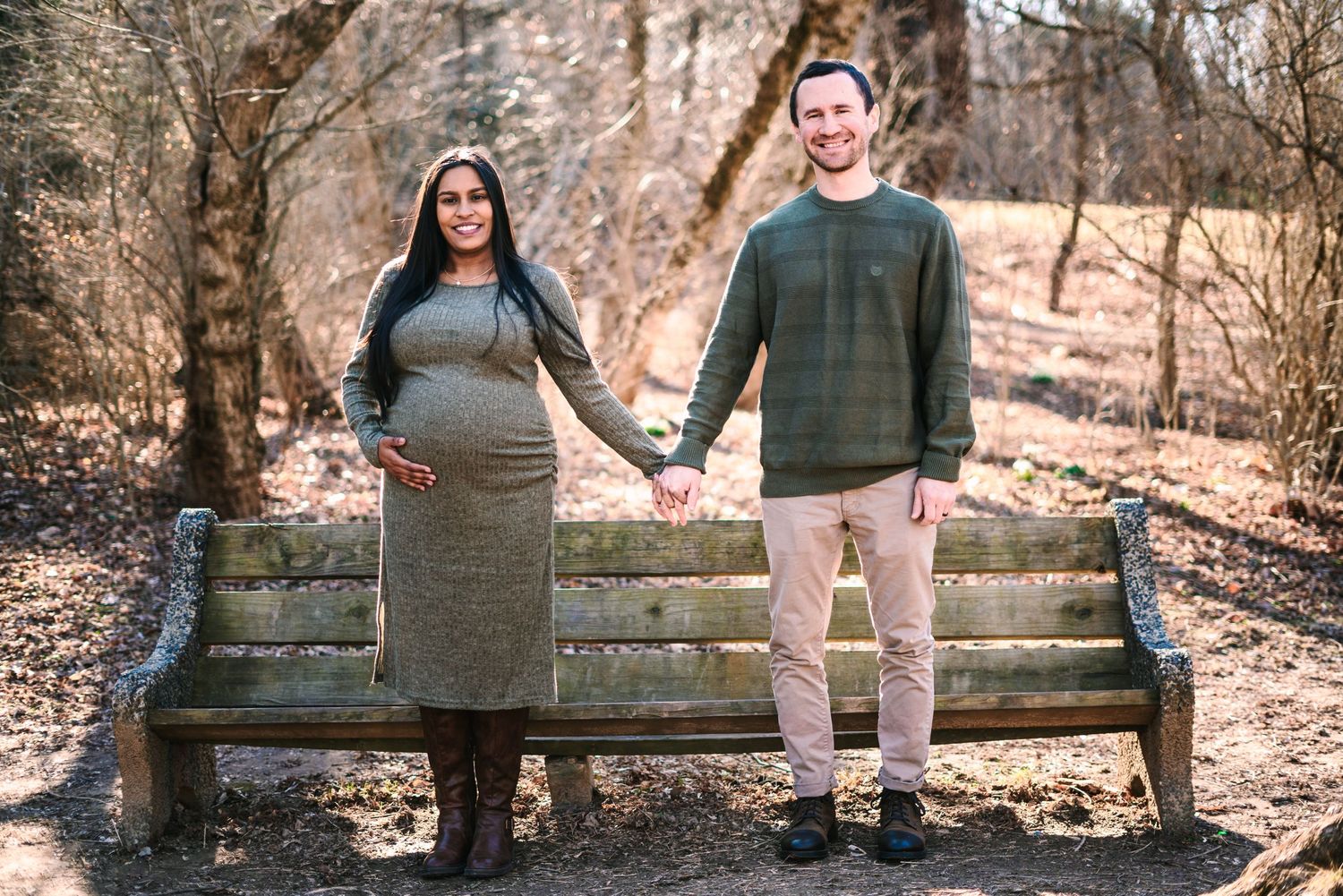 Pregnant couple holding hands in front of a wooden bench outdoors during a maternity photo shoot.