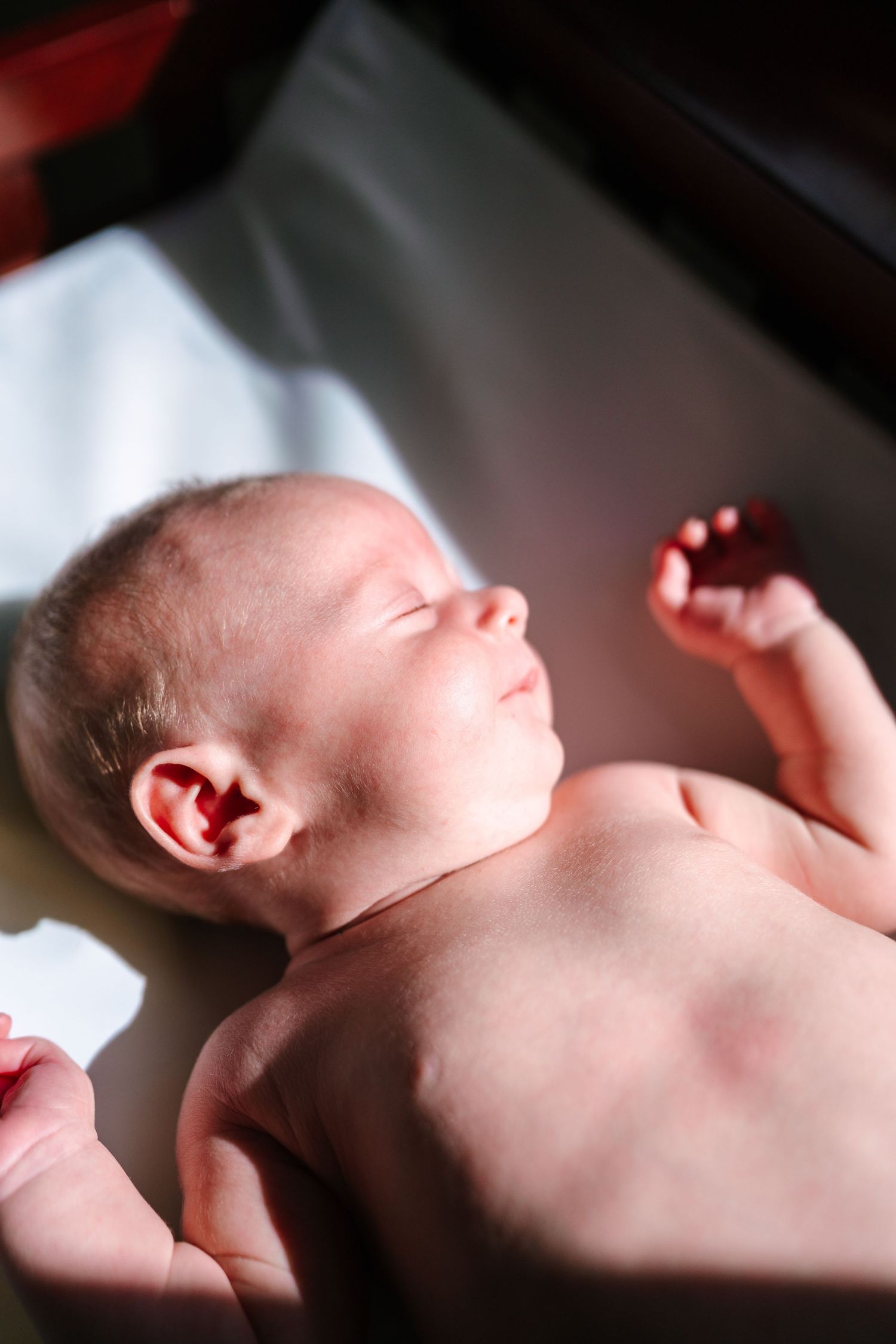 Newborn baby lying on white blanket with eyes closed, tiny hands raised in natural light.