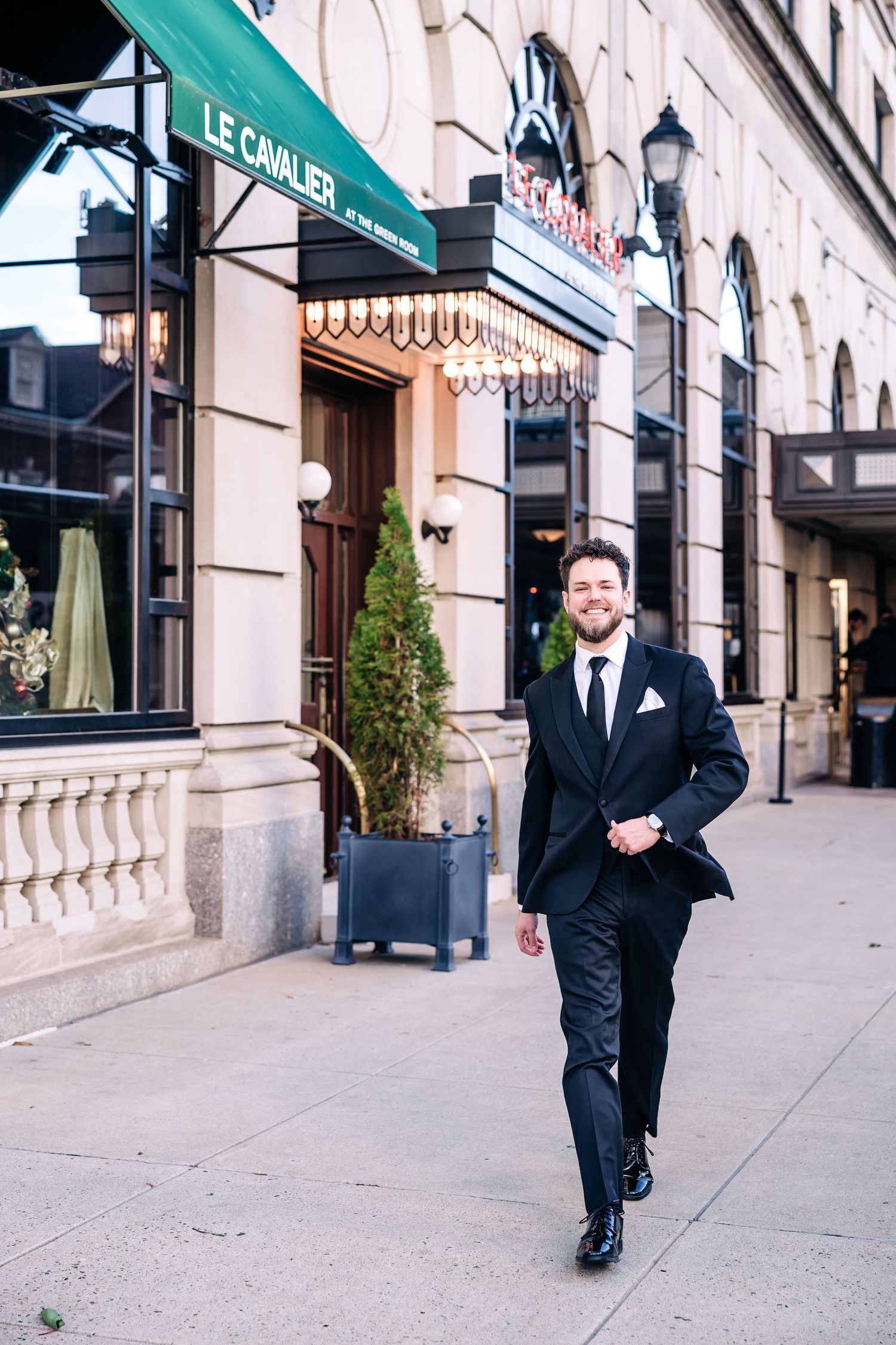 Smiling man in black tuxedo walking on sidewalk outside Le Cavalier restaurant in elegant urban setting Hotel DuPont wedding
