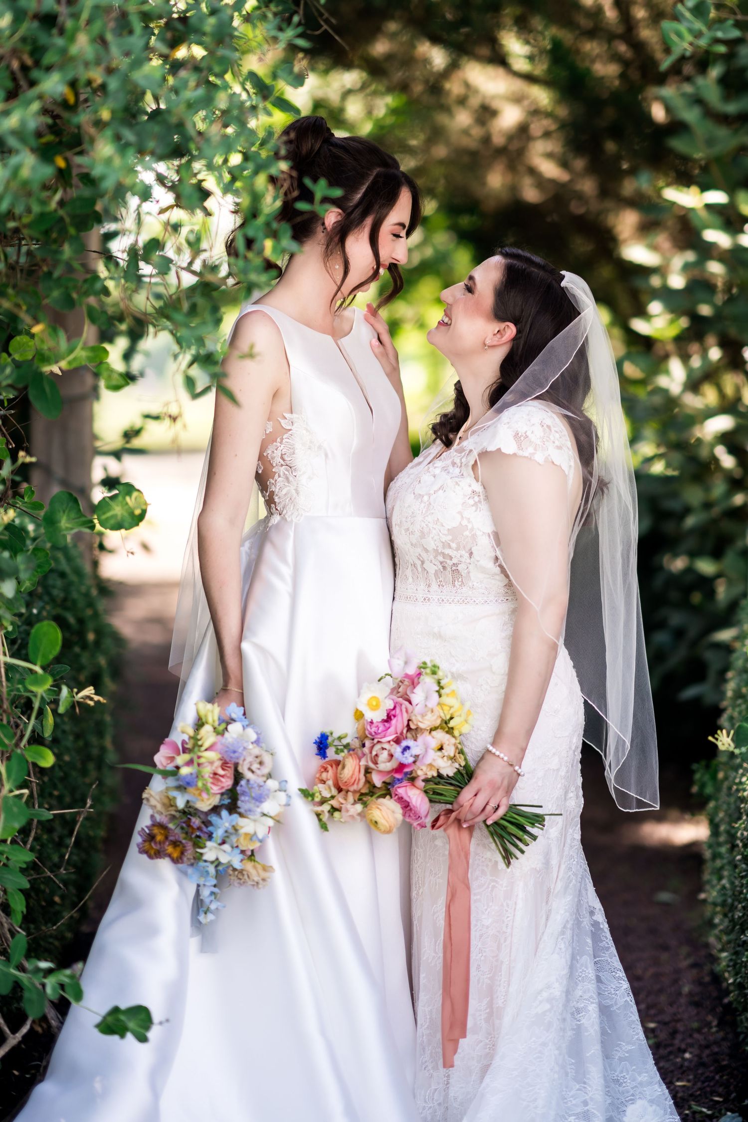 Fernbrook farms Philly Two brides in white wedding gowns holding colorful bouquets, sharing a romantic moment in a lush garden setting.