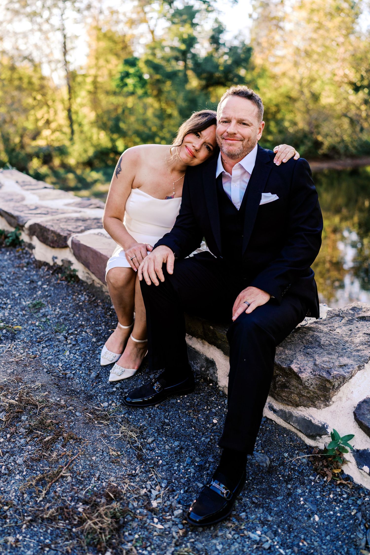 philadelphia wedding photography  A couple in wedding attire sits on a stone wall outdoors, smiling warmly, surrounded by lush greenery.
