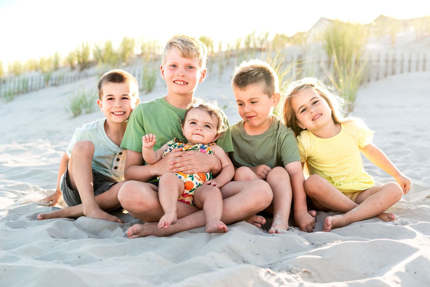 Five young children sitting together on sandy beach, smiling happily in colorful summer outfits with dunes in background.