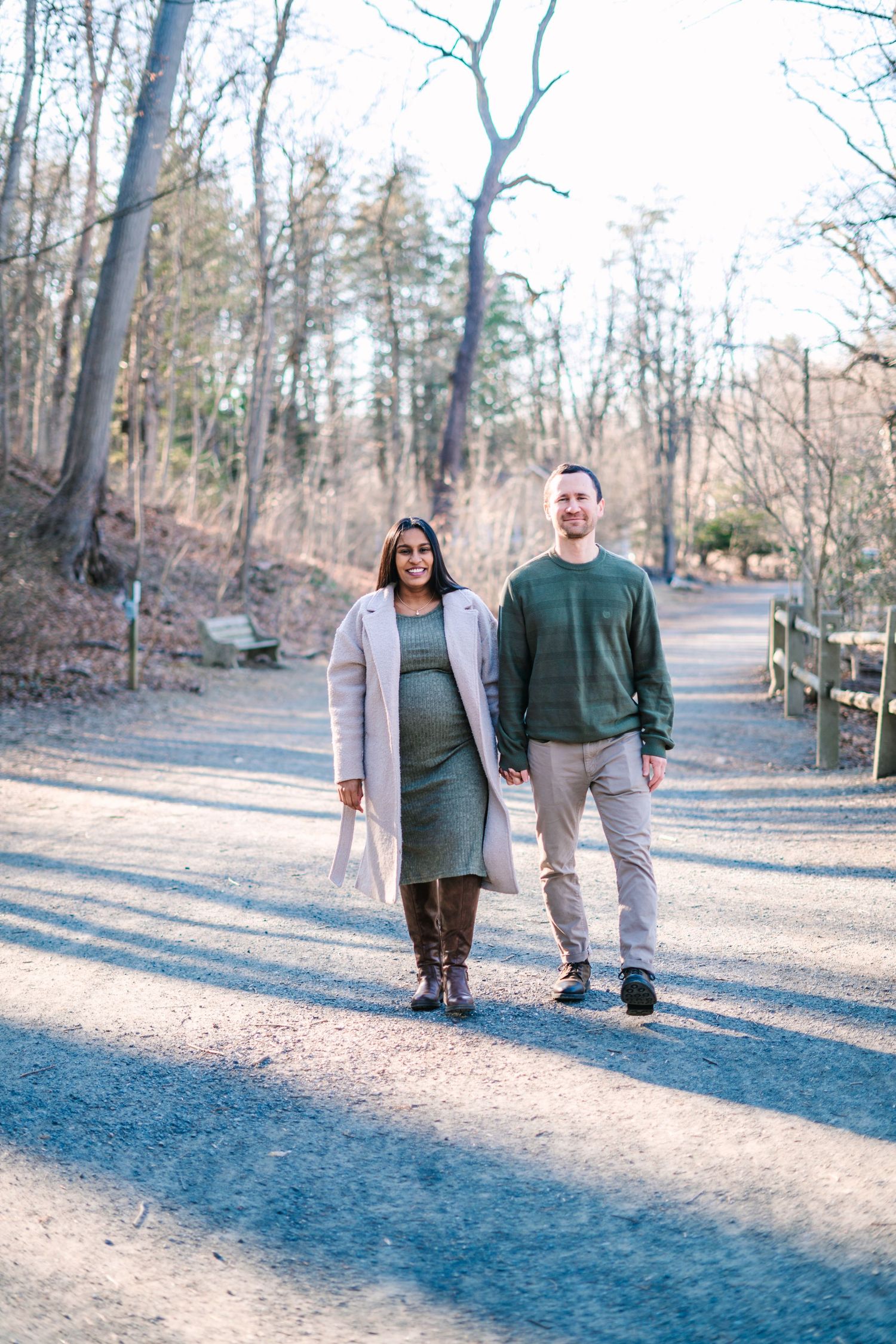 Pregnant couple walking hand-in-hand on a park path surrounded by bare trees in autumn.