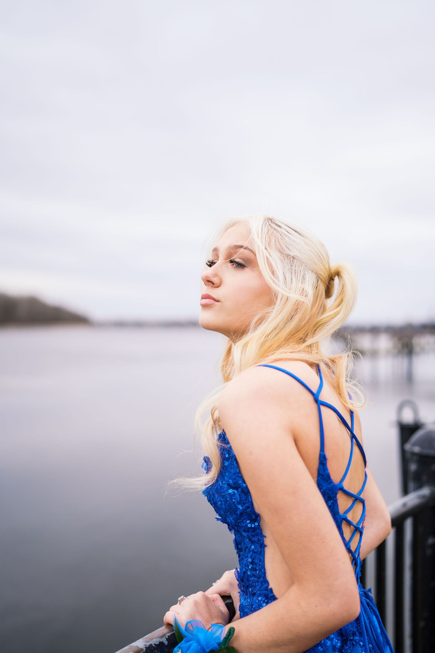 Blonde woman in blue sequin prom dress poses by waterfront railing at dusk.