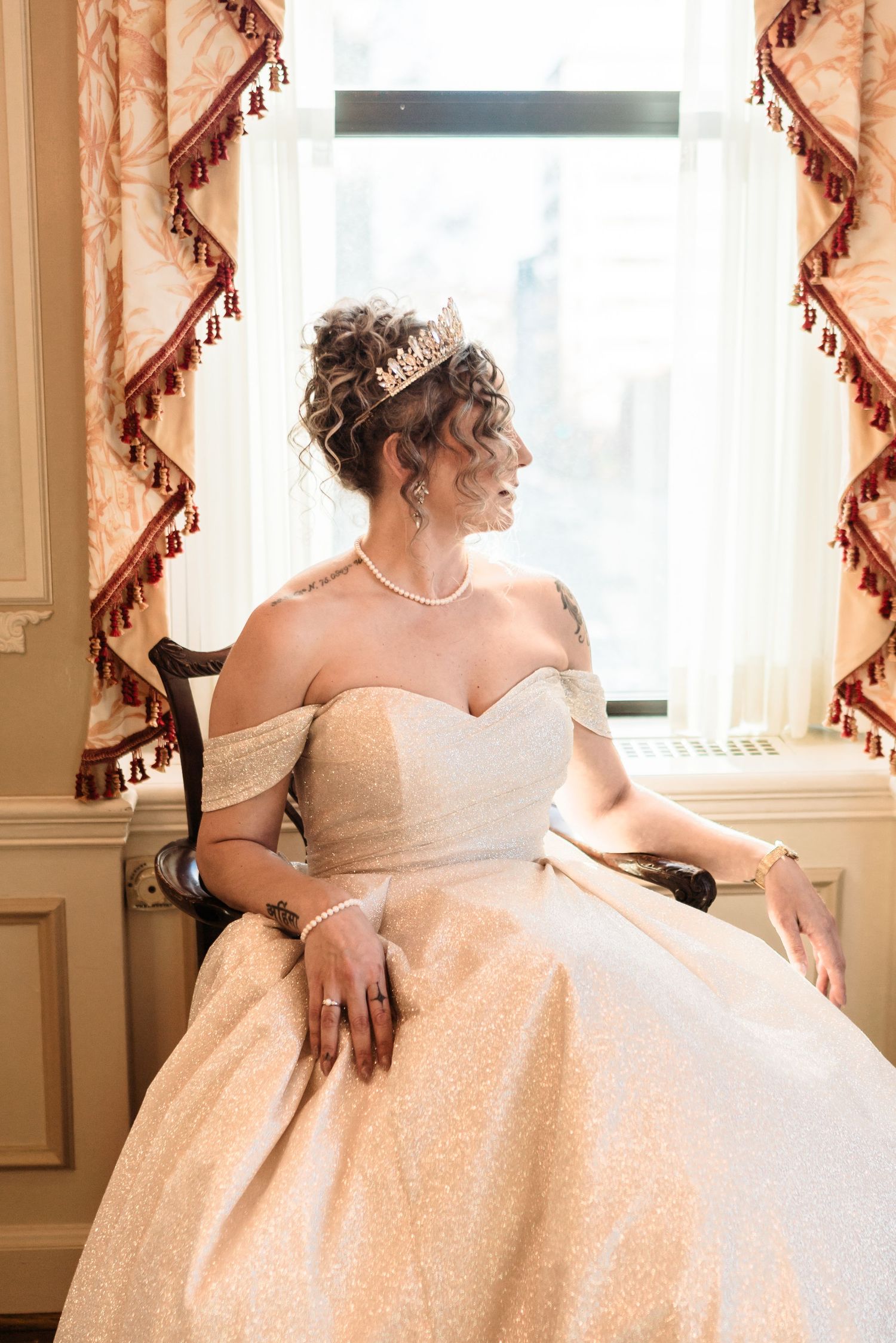 Bride in off-shoulder gown and tiara sits gracefully by a sunlit window with ornate curtains Hotel DuPont wedding Philadelphia photographer