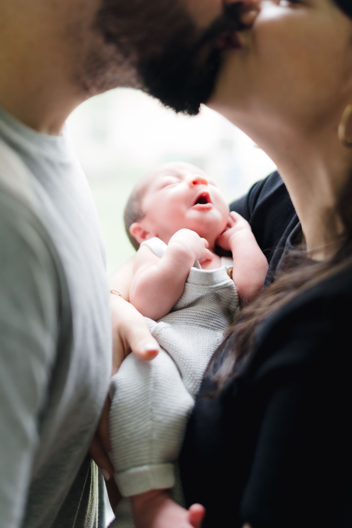 Parents kissing over their newborn baby held in arms, tender family moment with infant in gray onesie.