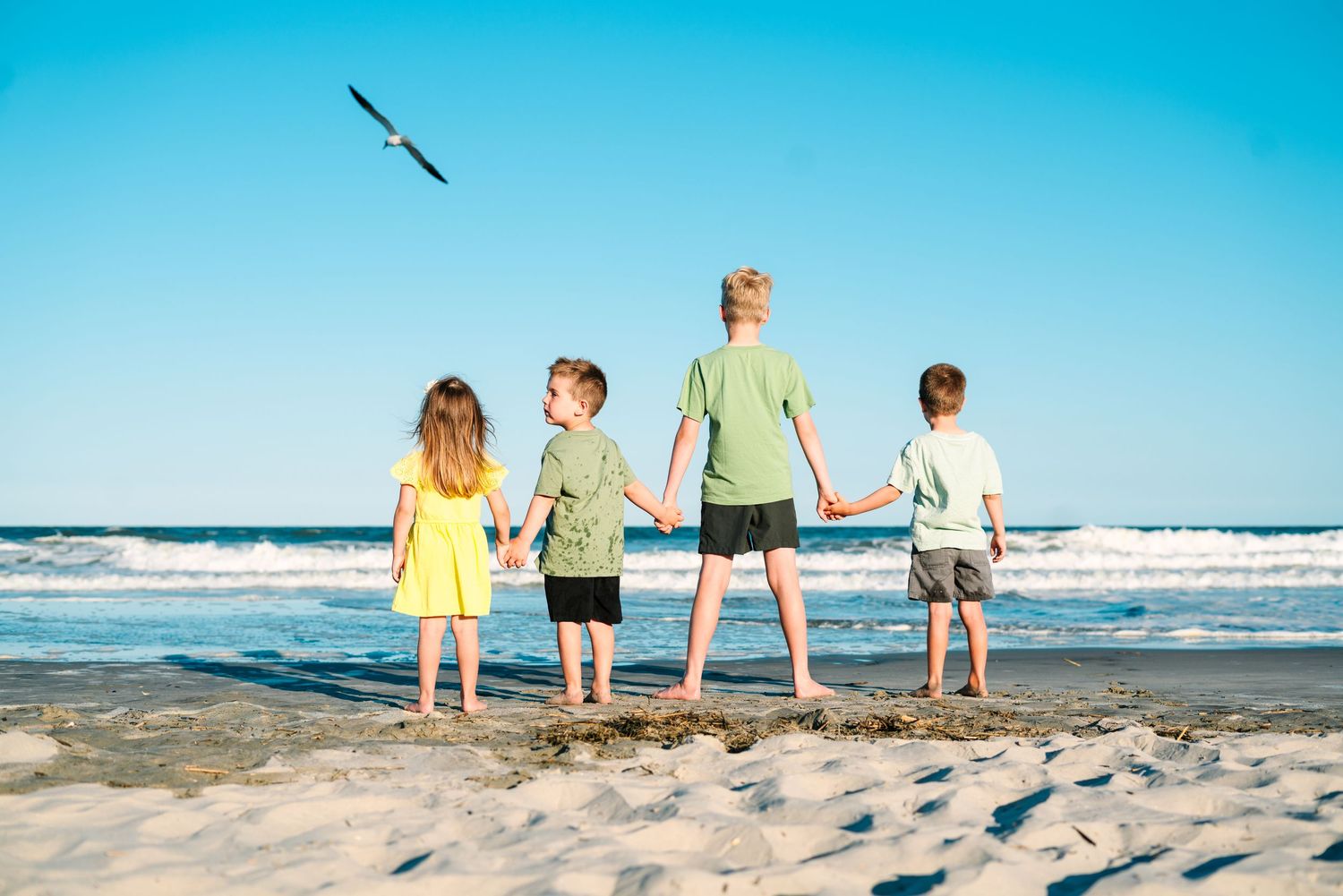 Four children holding hands on a sandy beach, watching ocean waves under a clear blue sky with a seagull flying overhead.