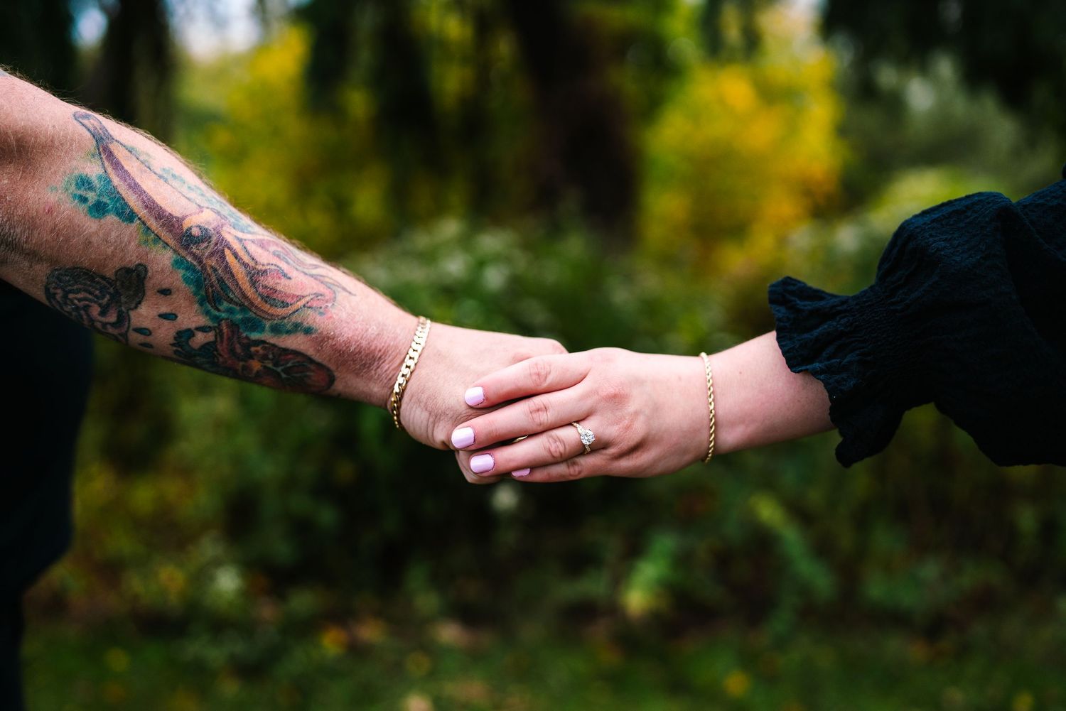 Tattooed arm and delicate hand with gold ring holding hands outdoors among autumn foliage.