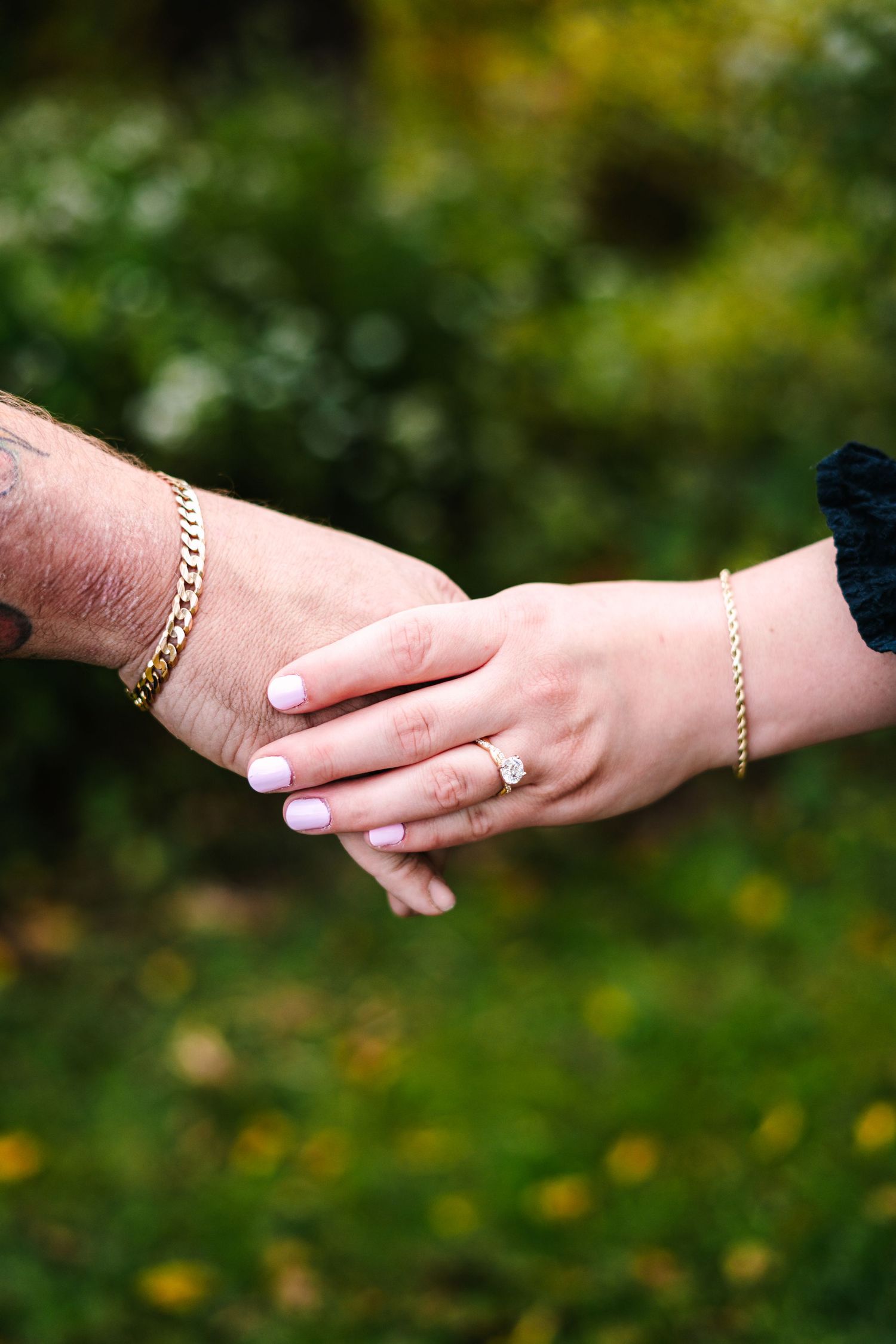 philadelphia wedding Close-up of couple holding hands showing engagement ring and gold bracelets outdoors.