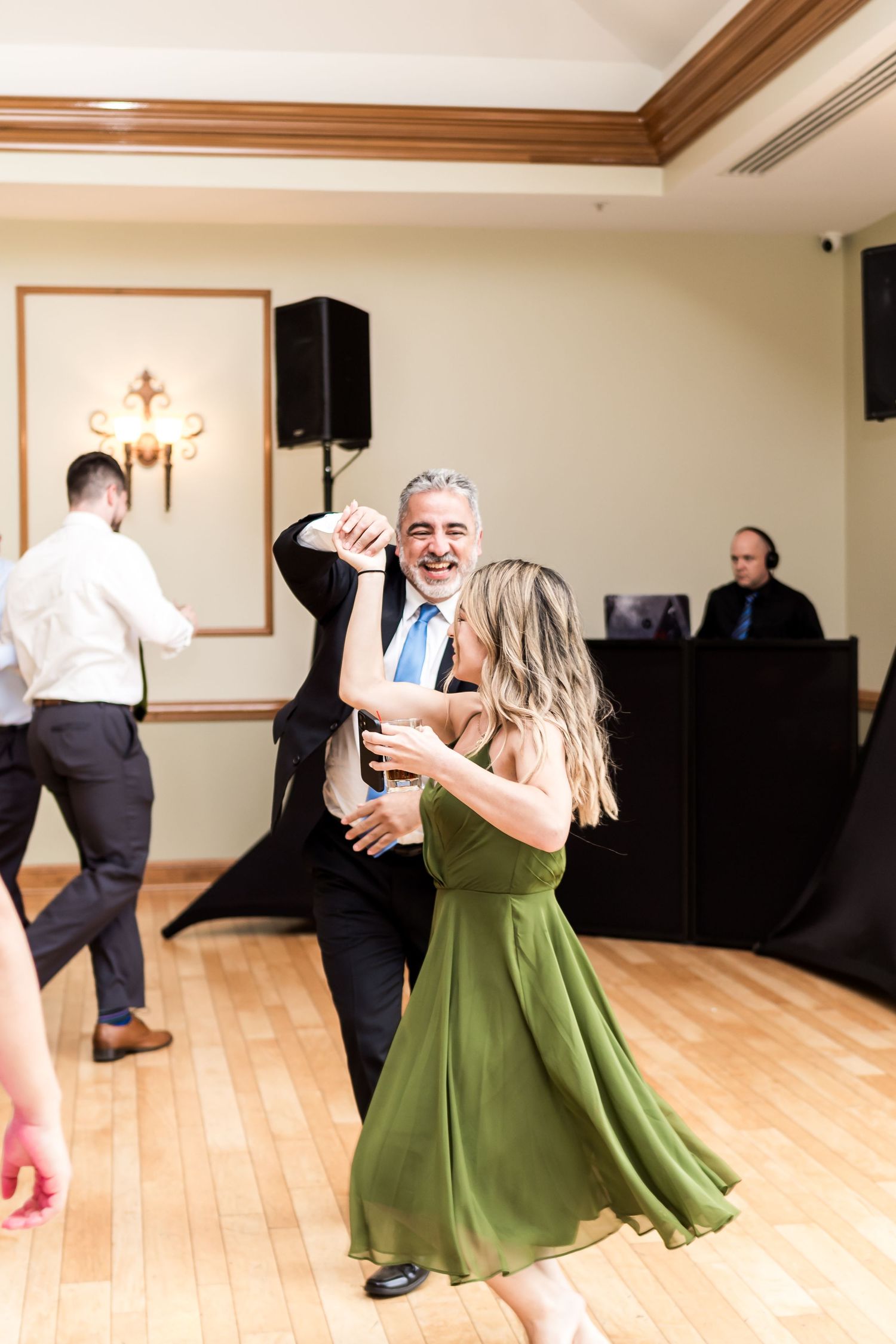 A couple dances joyfully at a wedding reception, the woman in a green dress, with a DJ and guests visible in the background.