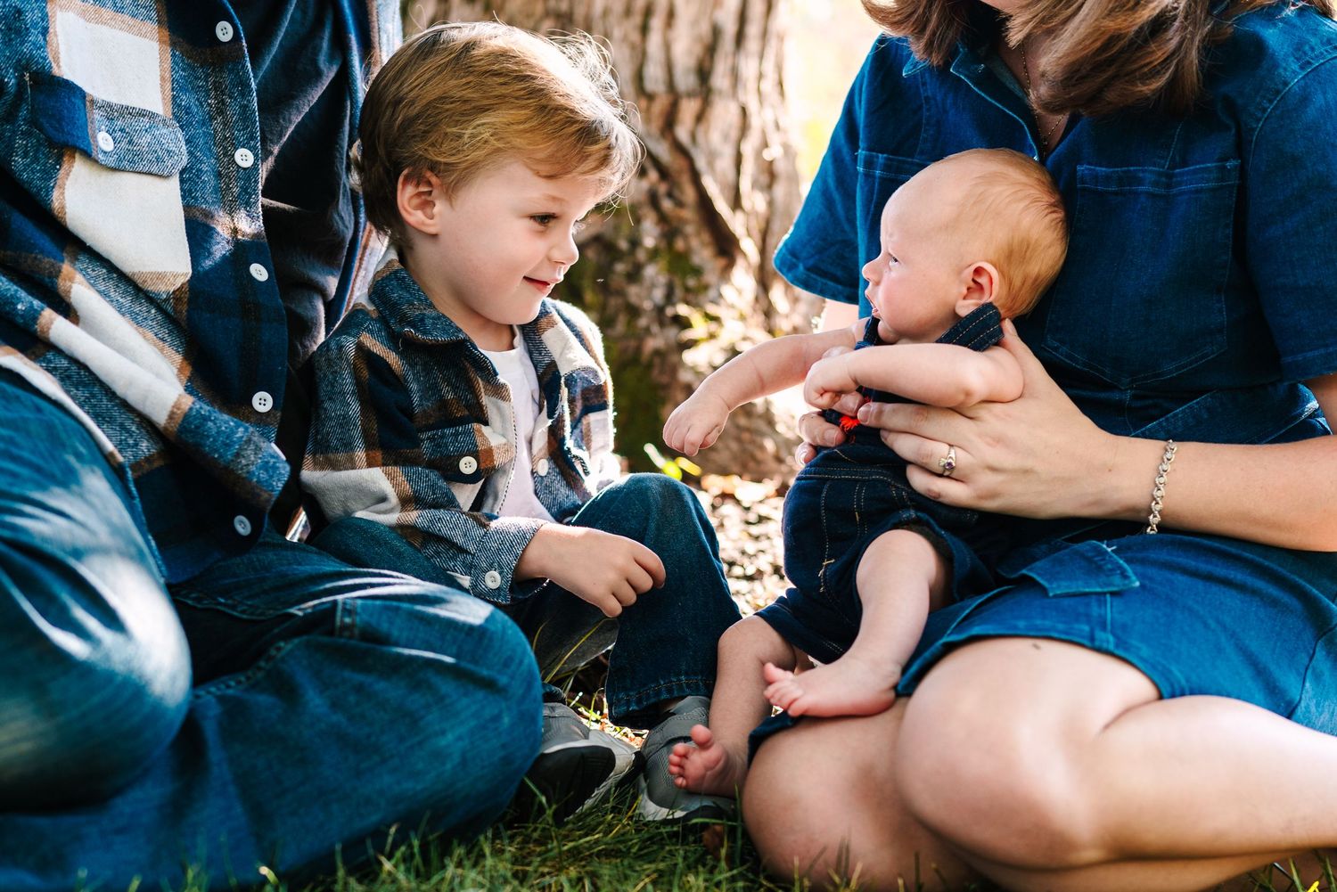 Family sitting outdoors with toddler boy and mother holding newborn baby near a tree.
