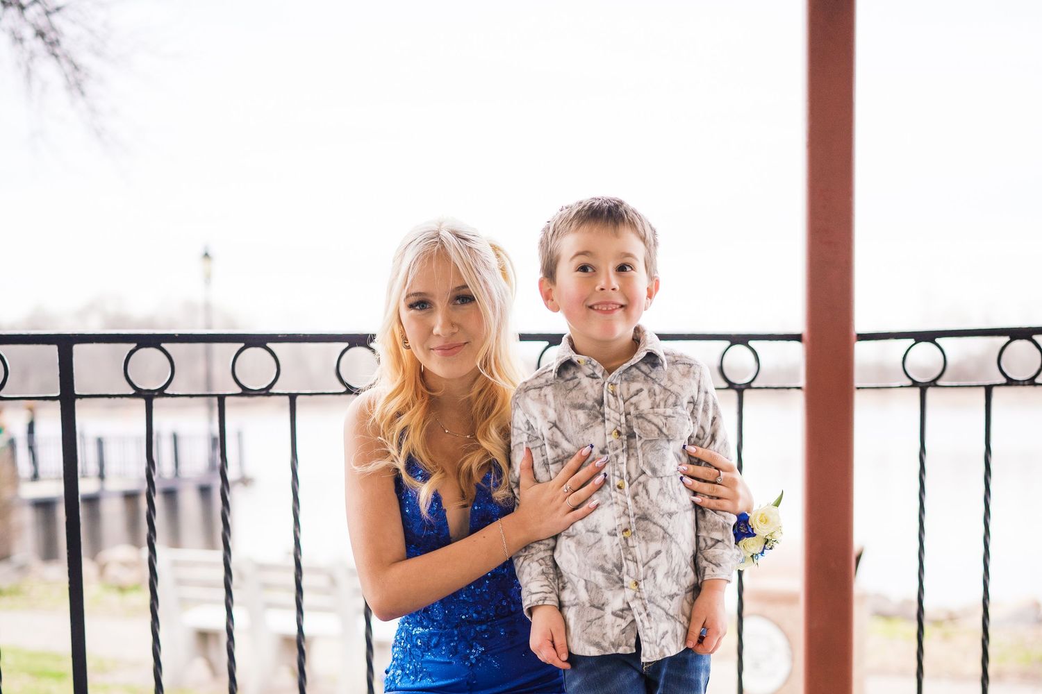 A young woman in a blue sequin dress poses with a smiling boy in a floral shirt on an outdoor balcony.