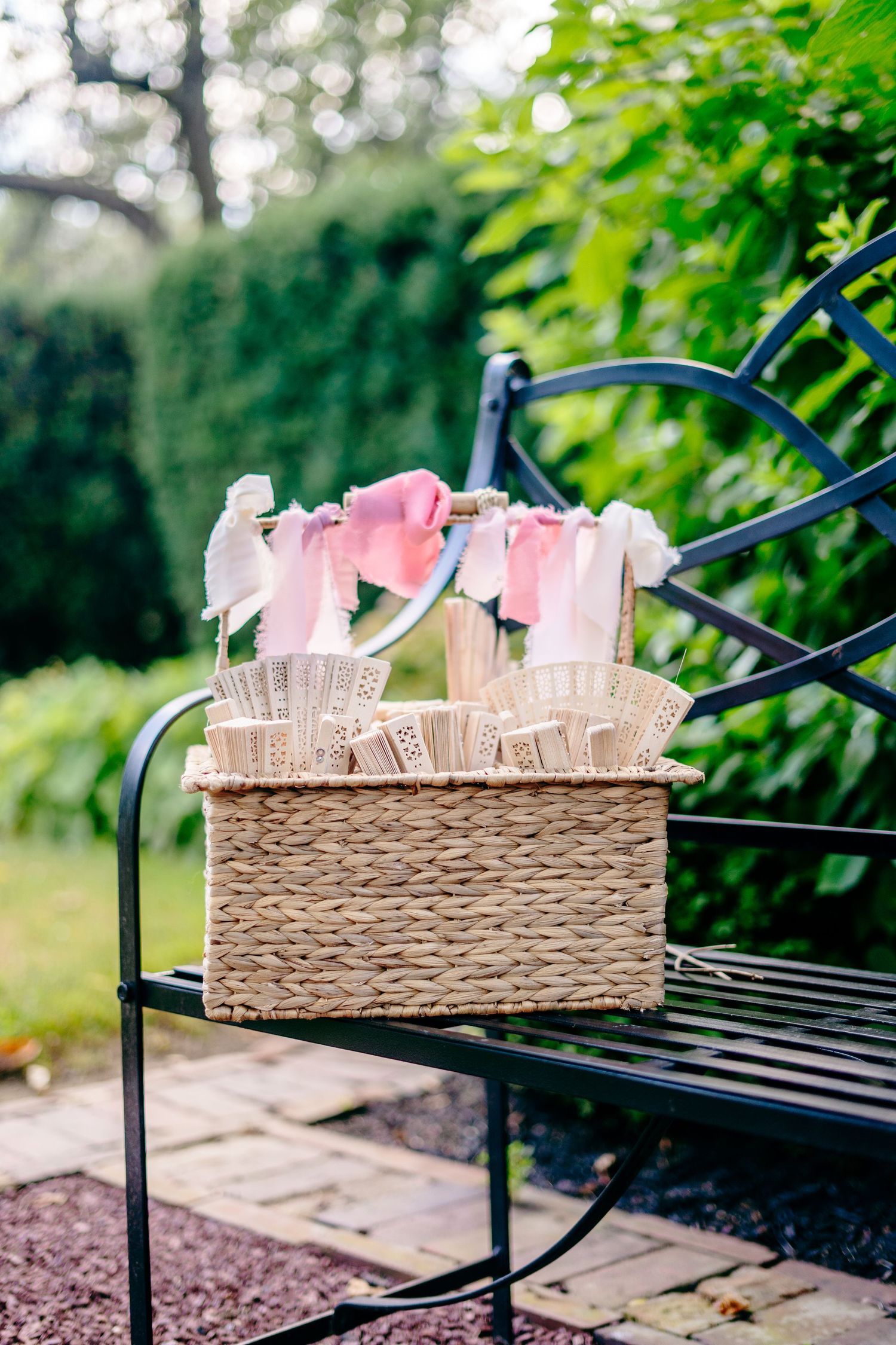Fernbrook farms Philly Wicker basket filled with decorative paper fans and pink ribbons displayed on a black metal garden bench outdoors.