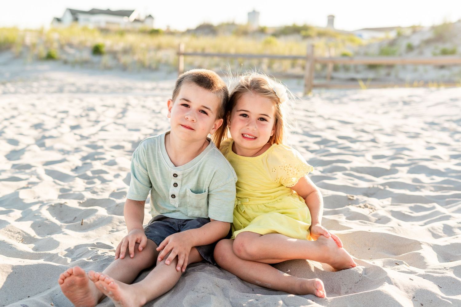 Two young children sitting together on a sandy beach, smiling, boy in green shirt and girl in yellow dress.