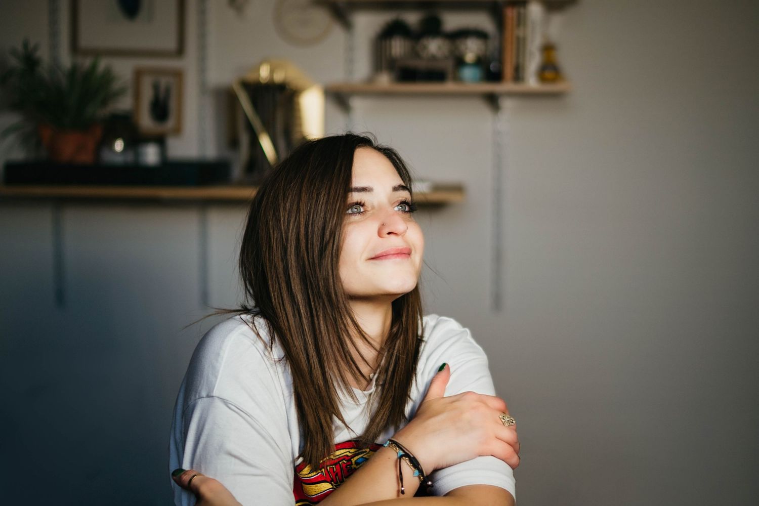 Young woman with long brown hair smiling dreamily, arms crossed, wearing a white t-shirt in a cozy home setting.