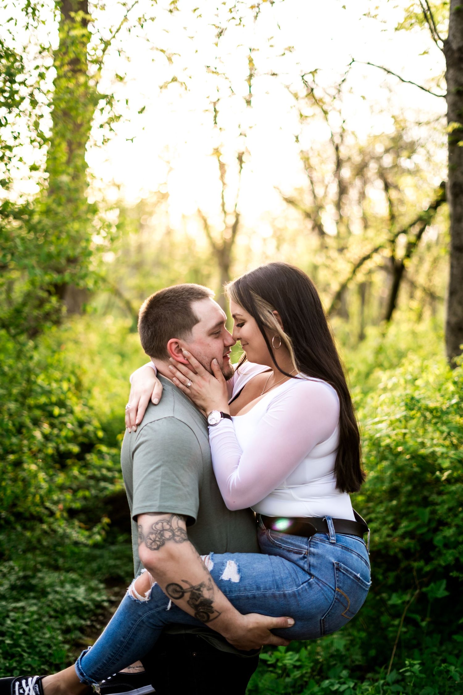 Couple sharing a romantic moment outdoors, man lifting woman in sunlit forest during engagement photo session.