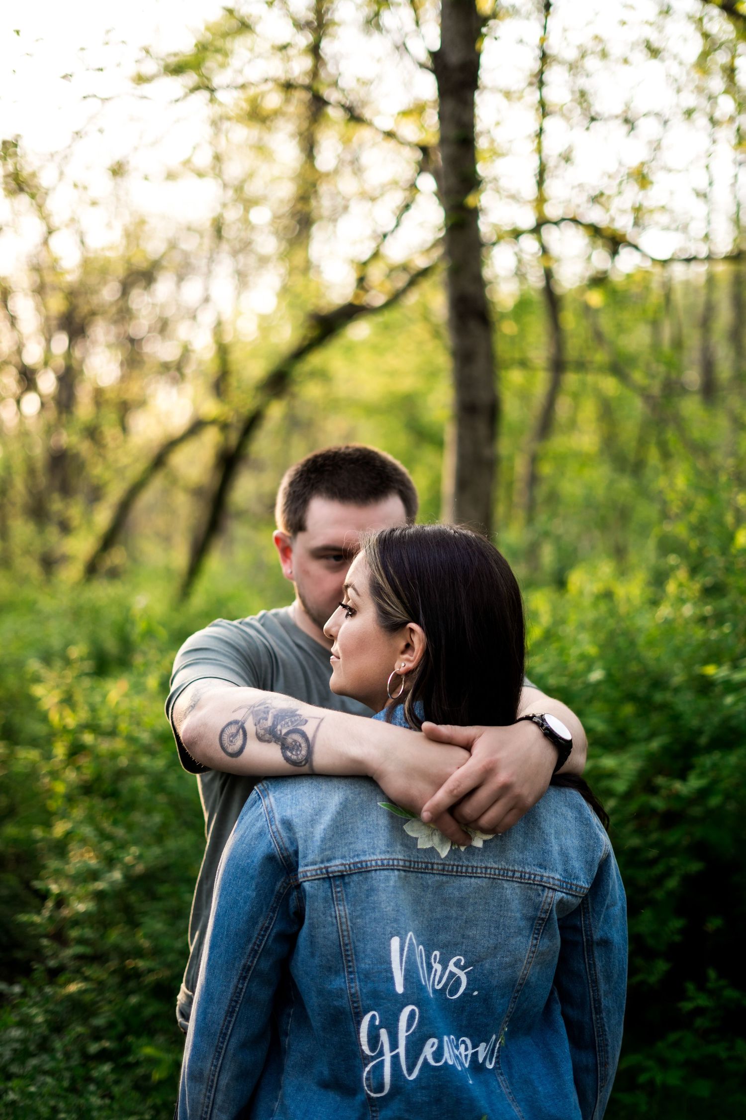 Couple embracing outdoors, woman wearing custom denim jacket reading Mrs. Glenn, surrounded by lush green forest.