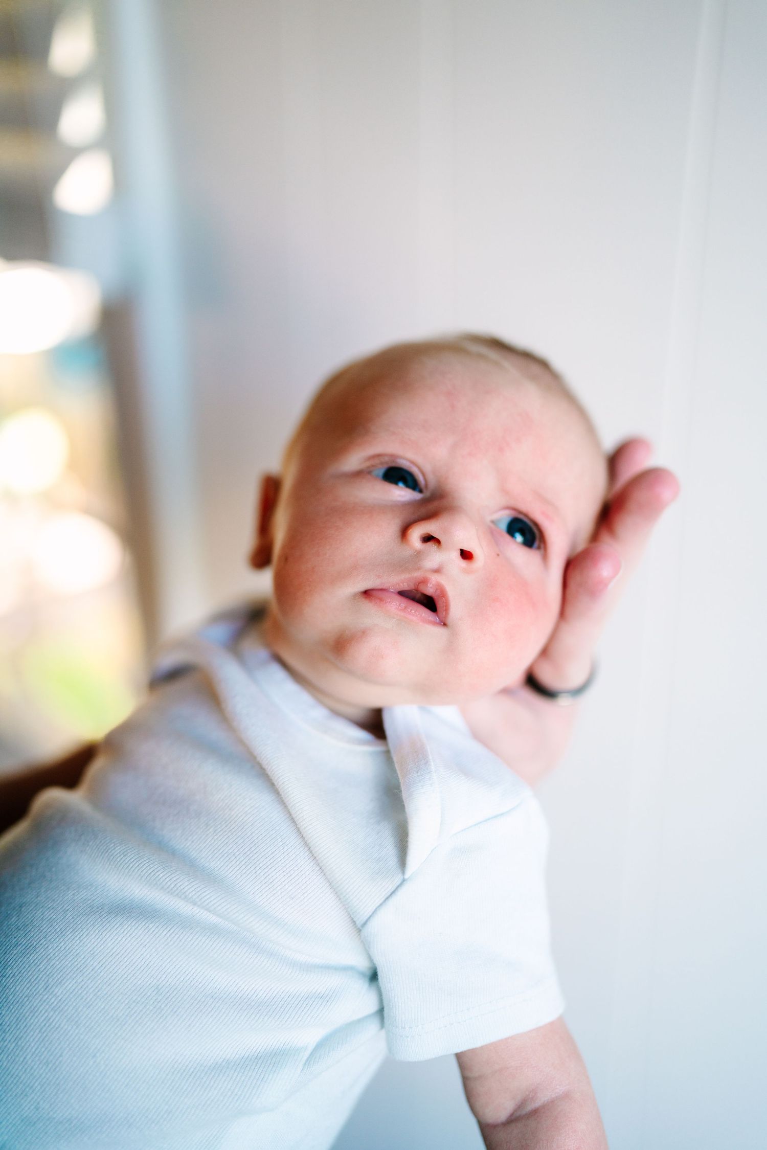 Newborn baby in white onesie with mittens gazing upward, held by a gentle hand near a bright window.