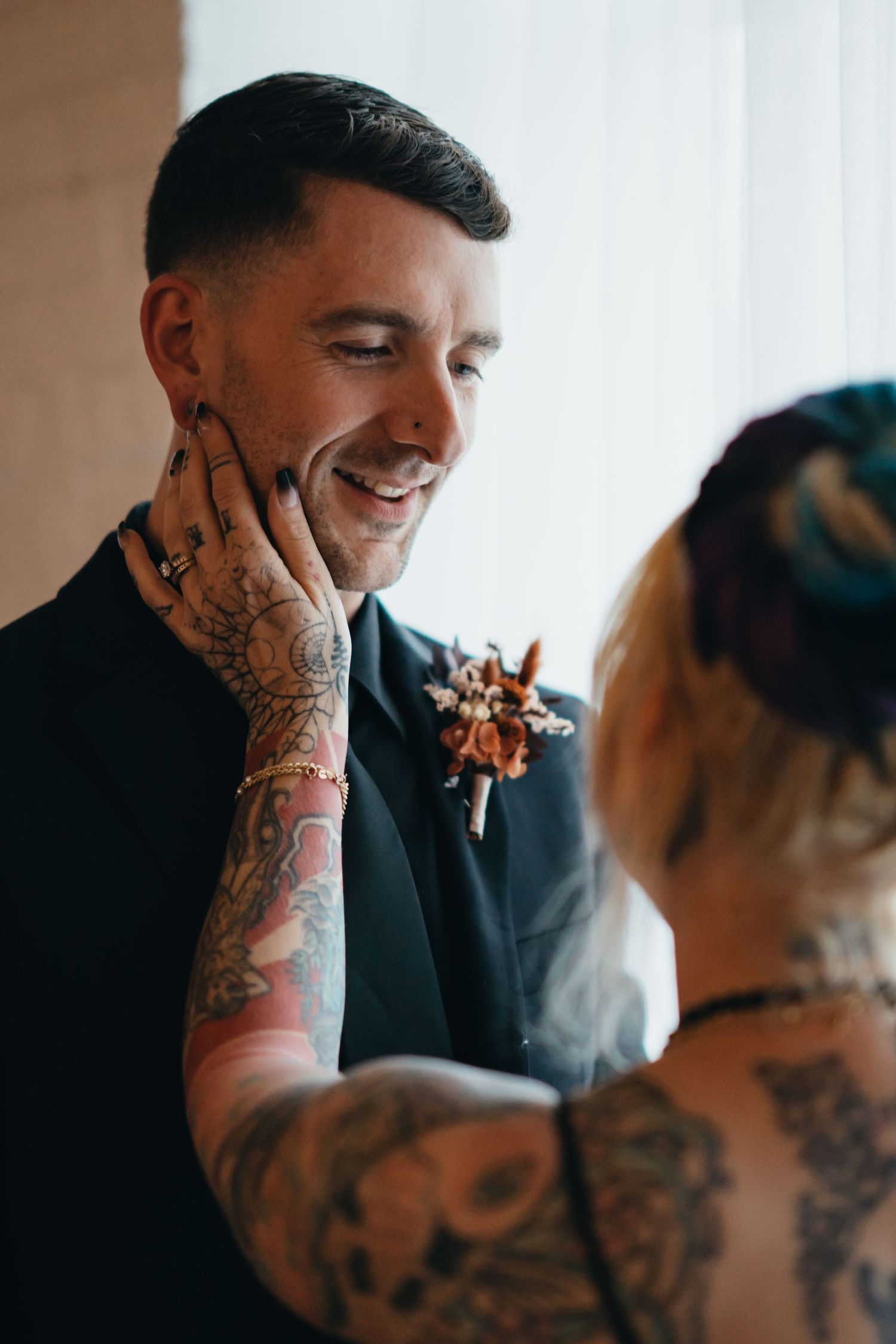 Smiling groom in black suit with boutonniere, tattooed bride tenderly touching his face at wedding ceremony.