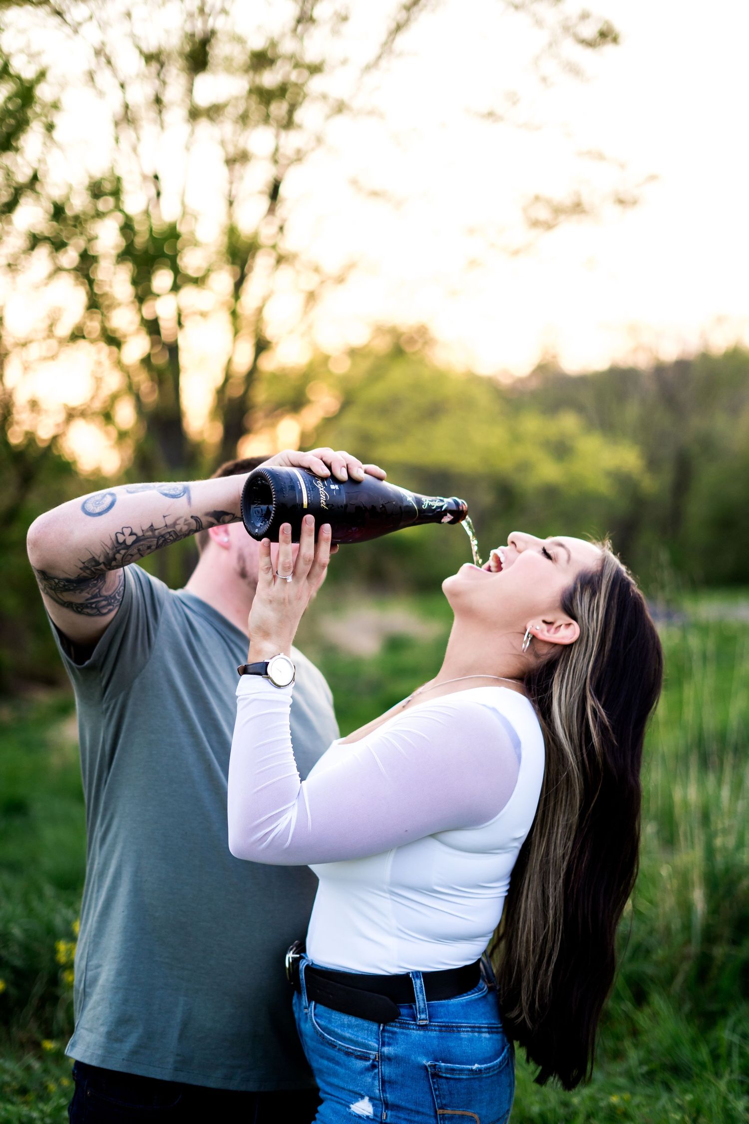 Couple outdoors at sunset, man pouring champagne into laughing woman's mouth, green trees background.