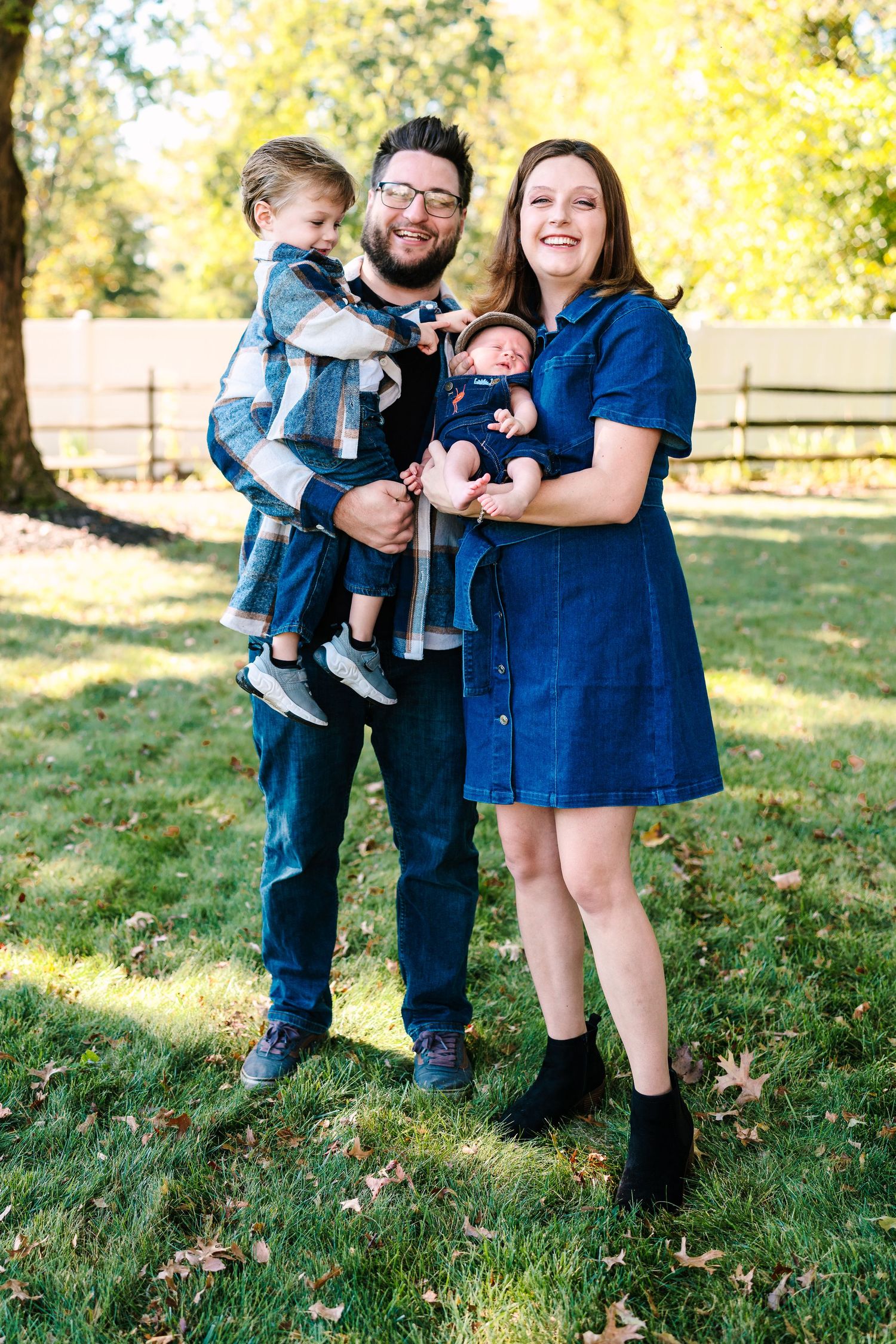 Happy family of four posing outdoors in fall, dad holding toddler and mom carrying newborn in blue carrier.