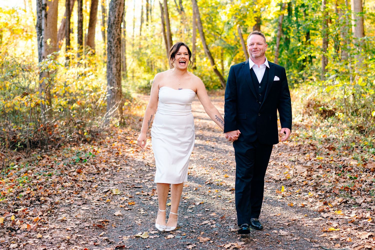 philadelphia wedding photography  Happy couple in wedding attire walking hand-in-hand on a forest path surrounded by autumn foliage.