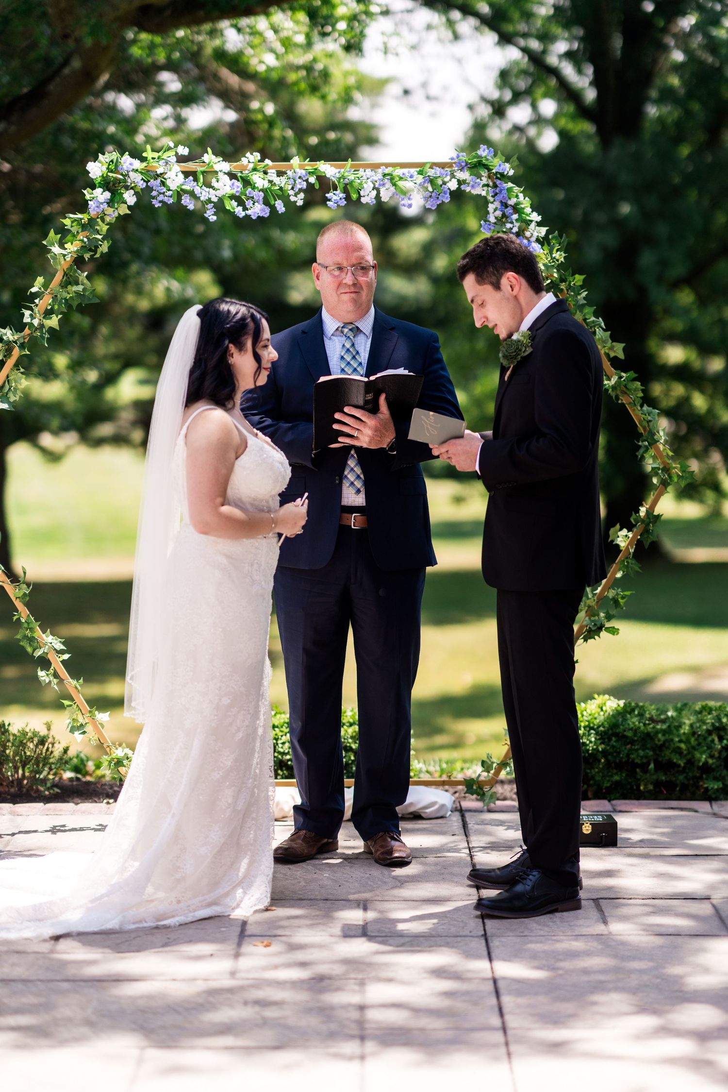 philadelphia wedding Bride and groom exchange vows outdoors under a floral arch with an officiant during a sunny wedding ceremony.