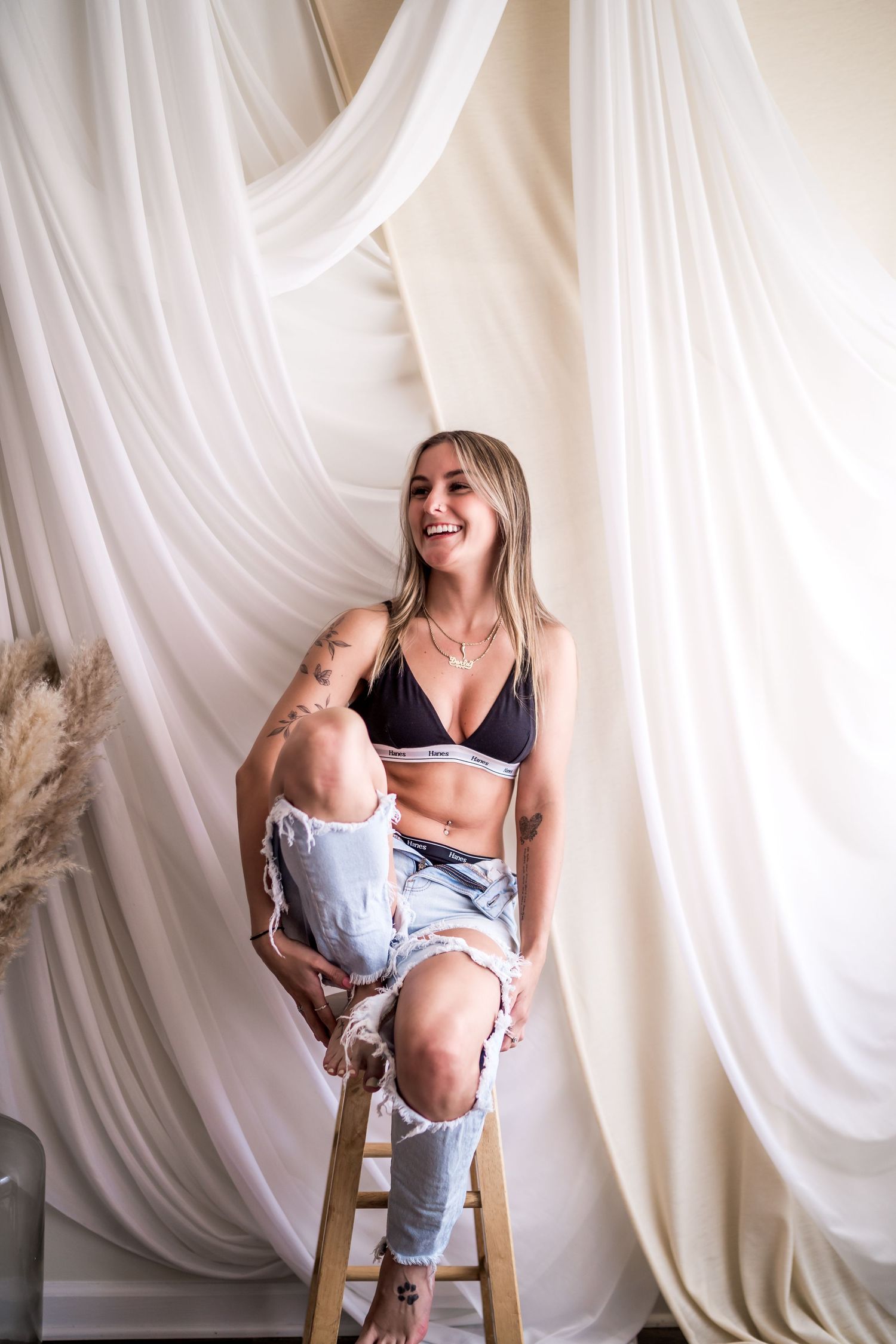 Smiling blonde woman in black bralette and ripped jeans sitting on wooden stool against flowing white curtain backdrop.
