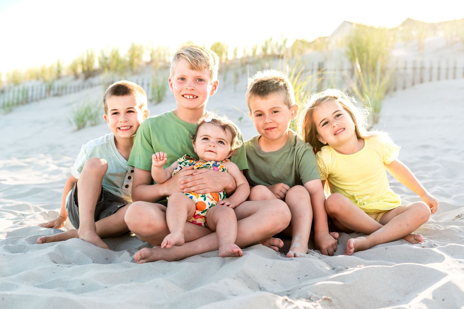 Five children sitting together on sandy beach, smiling happily, wearing colorful summer outfits with dunes in background.