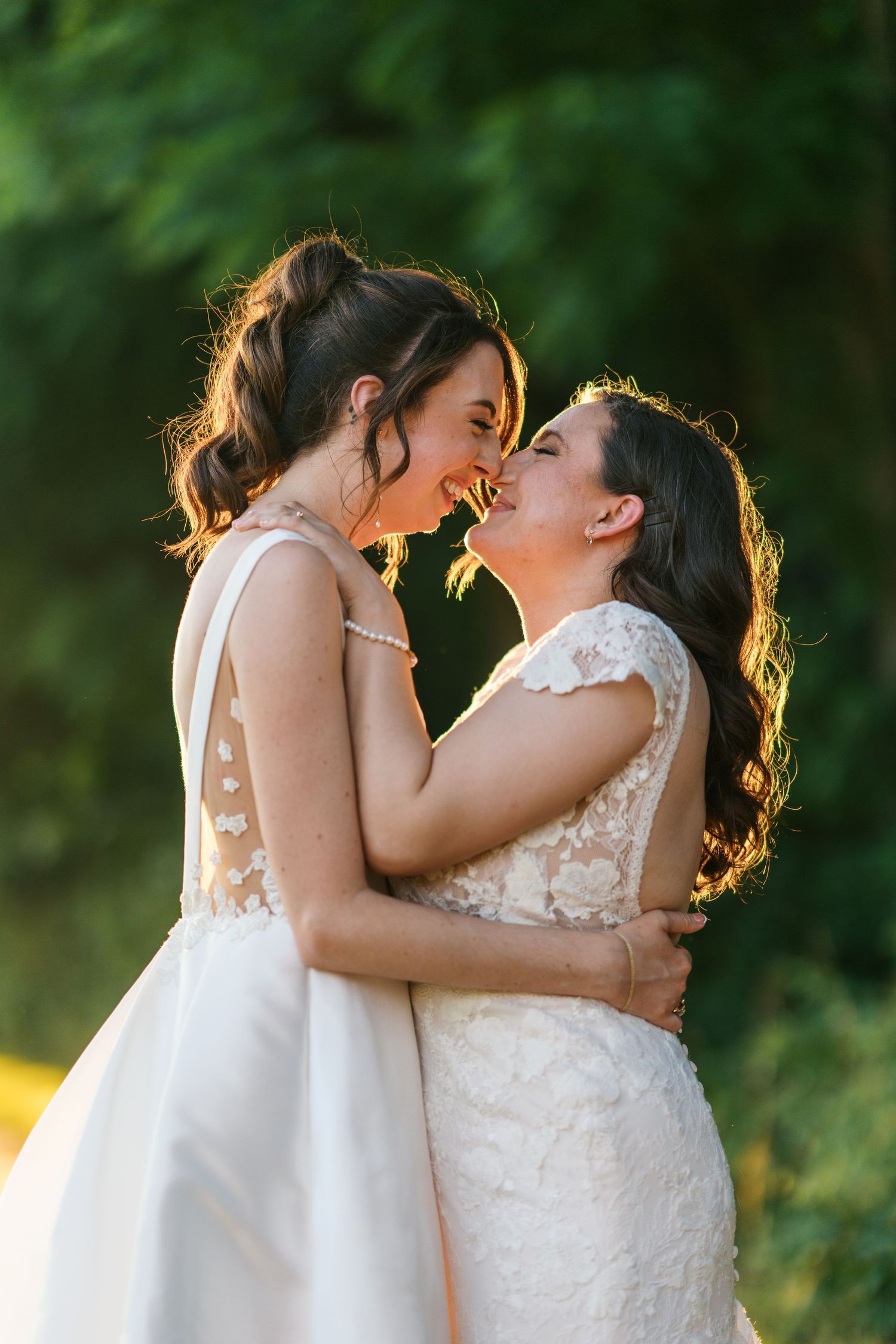 Fernbrook farms Philly wedding brides in white wedding gowns embrace and laugh together outdoors with lush green trees in the background.