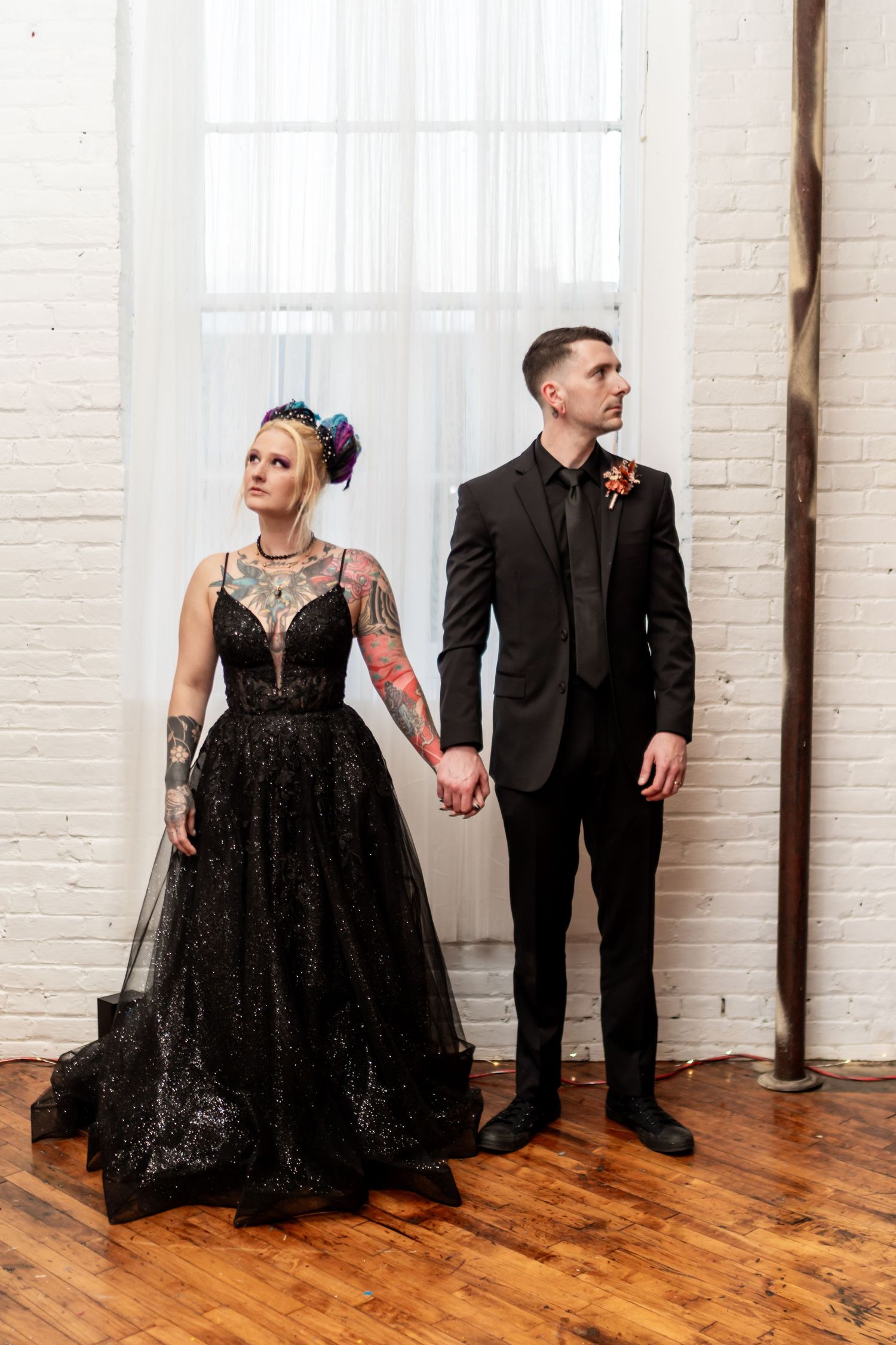 Couple in matching black wedding attire holding hands, bride in black tulle gown with tattoos, groom in black suit.