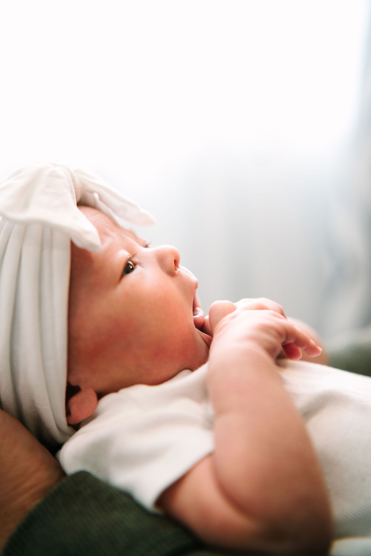 A newborn baby wearing a white bow headband lies peacefully, gazing upward with tiny hands clasped together.