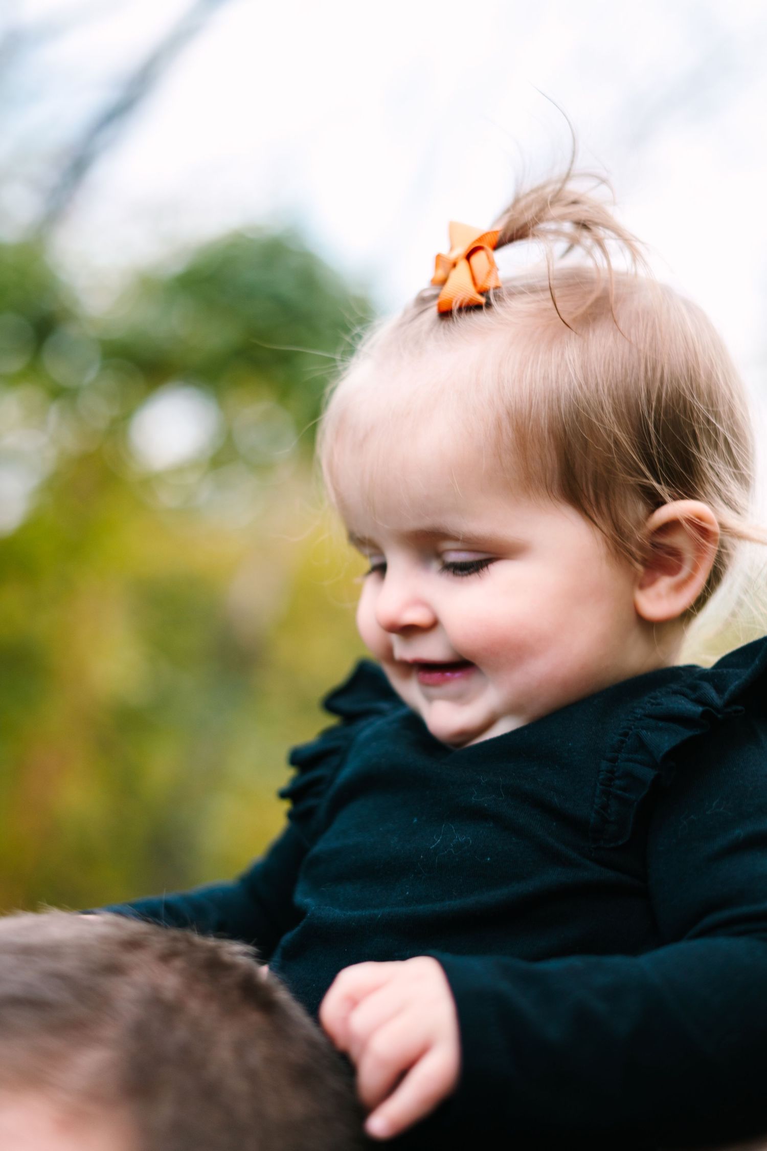 philadelphia wedding photography Smiling baby girl wearing an orange bow and dark green dress being held outdoors in autumn.