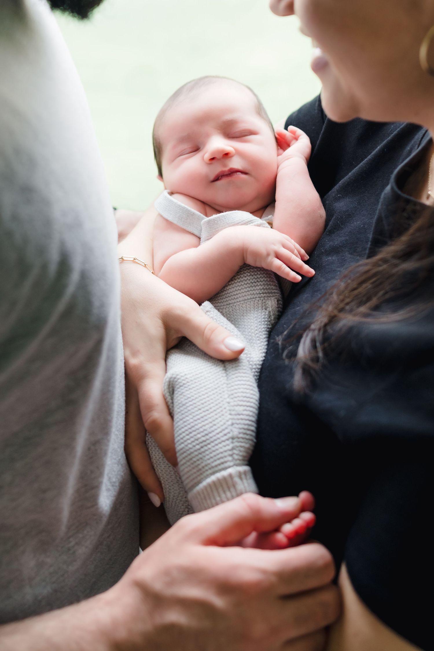 Parents cradling their sleeping newborn baby dressed in a gray onesie, close-up family portrait.
