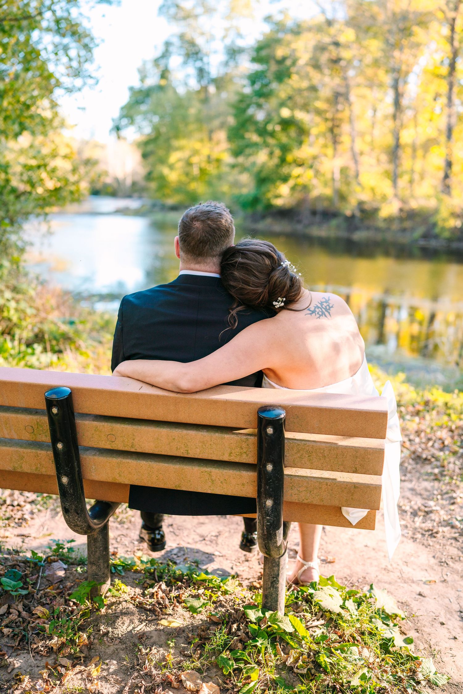 philadelphia wedding photography  Newlywed couple sitting on a wooden bench by a river, embracing and enjoying autumn scenery.