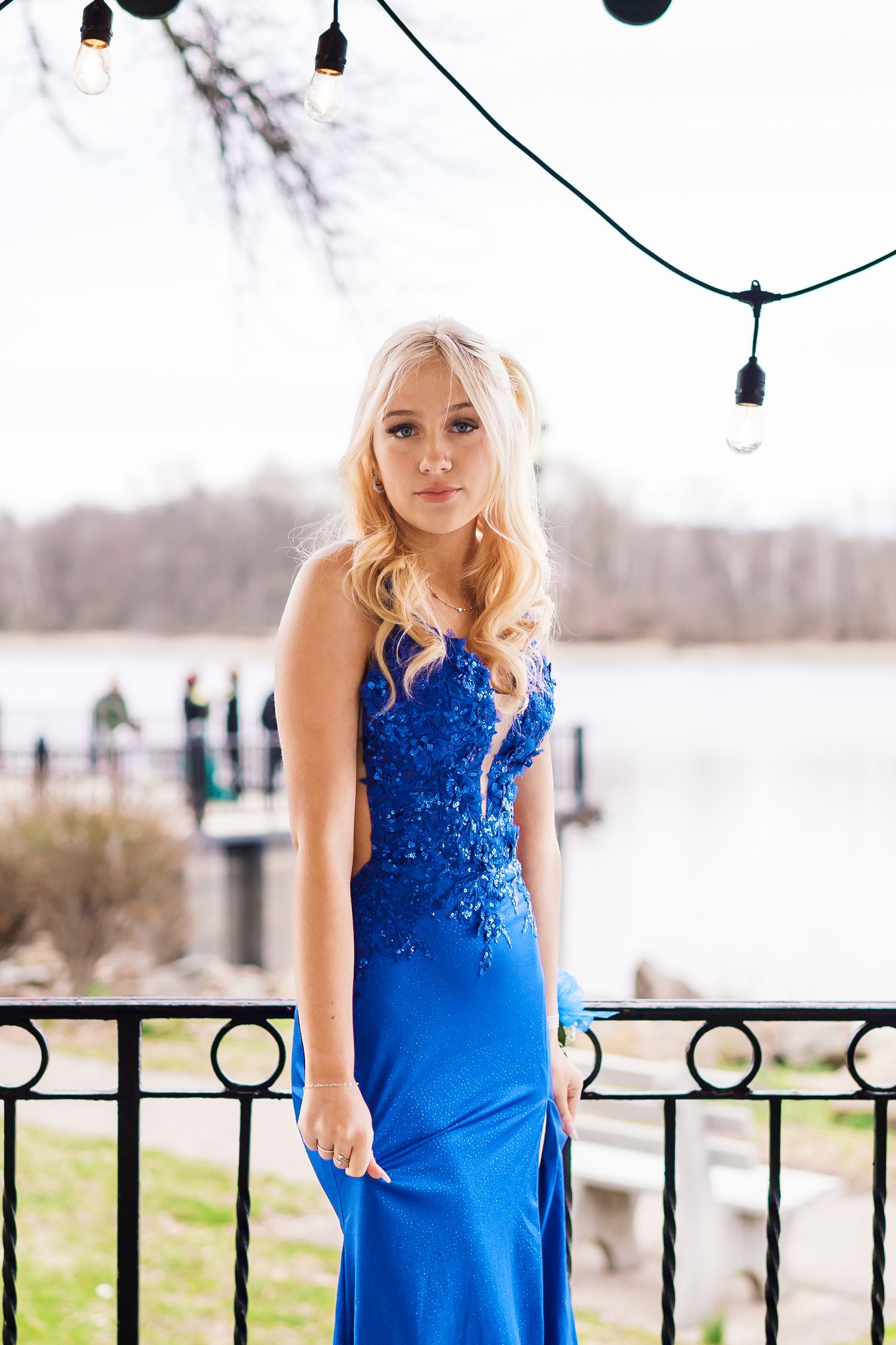 Blonde woman in elegant royal blue sequined formal gown posing by waterfront railing with string lights overhead.