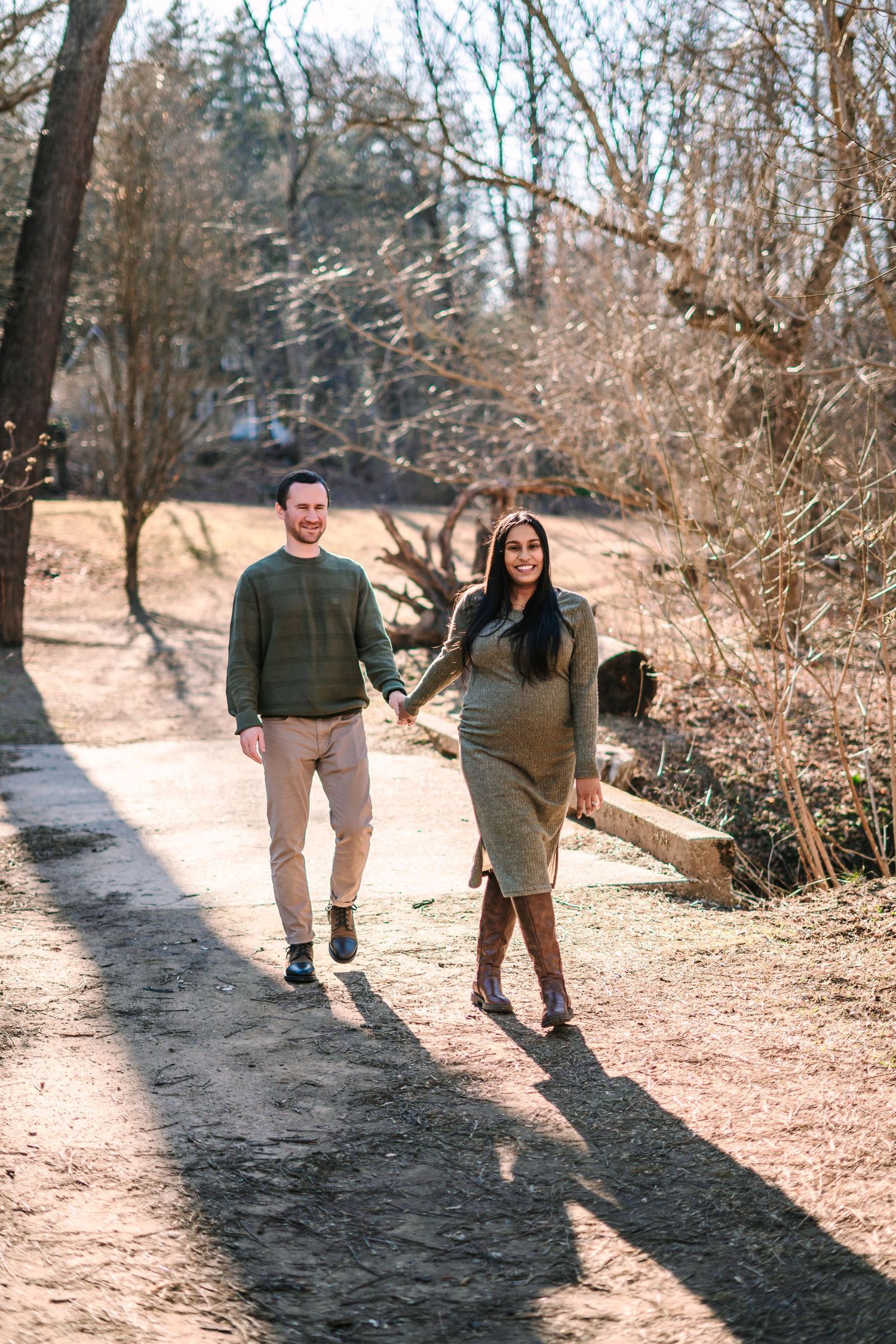 Pregnant couple holding hands walking on a sunny path surrounded by bare trees in a park.